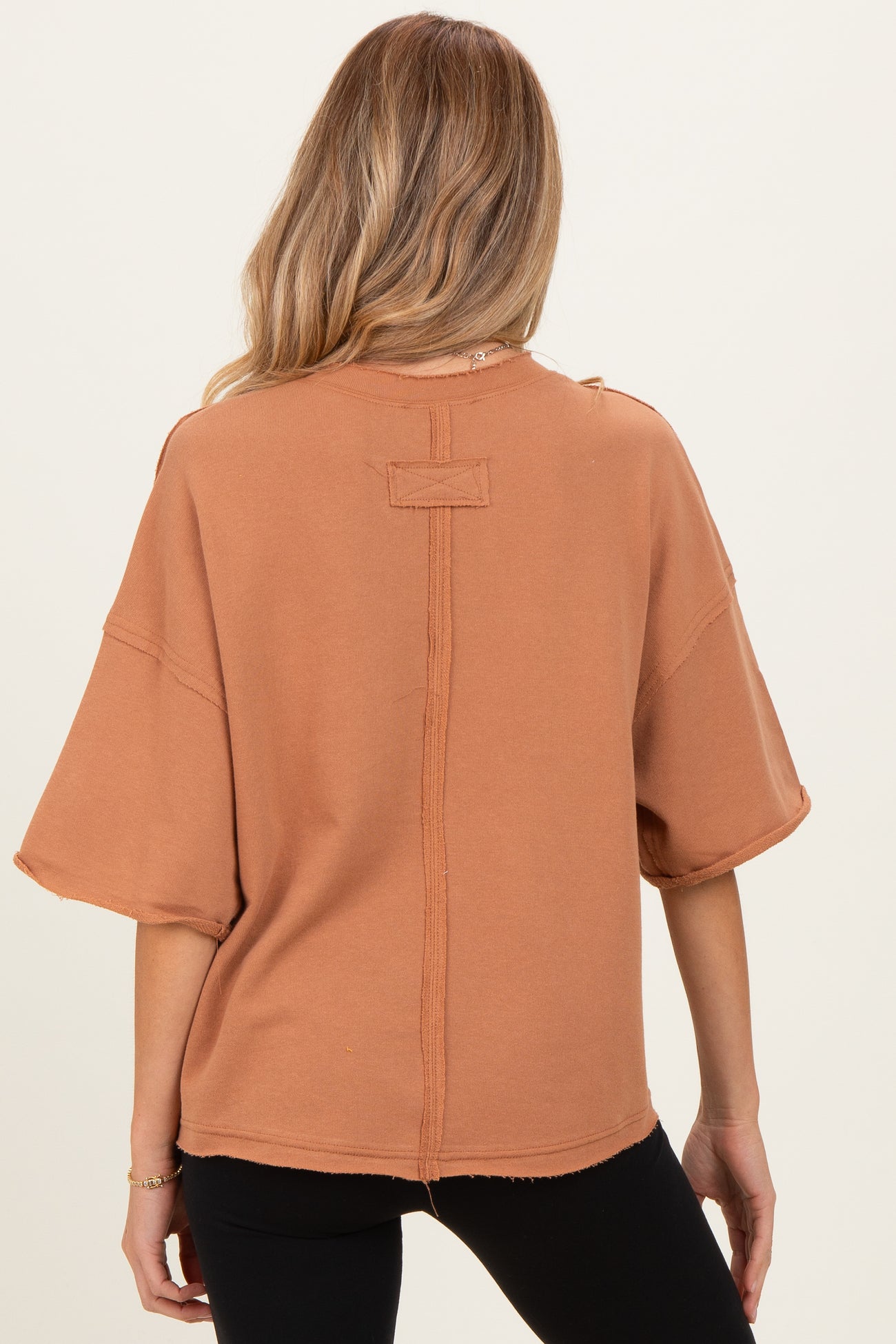 woman wearing a rust-colored oversized short sleeve maternity top with raw hem details, showing the back view