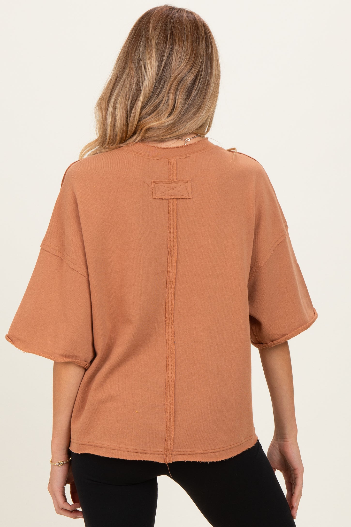 woman wearing a rust-colored oversized short sleeve maternity top with raw hem details, showing the back view