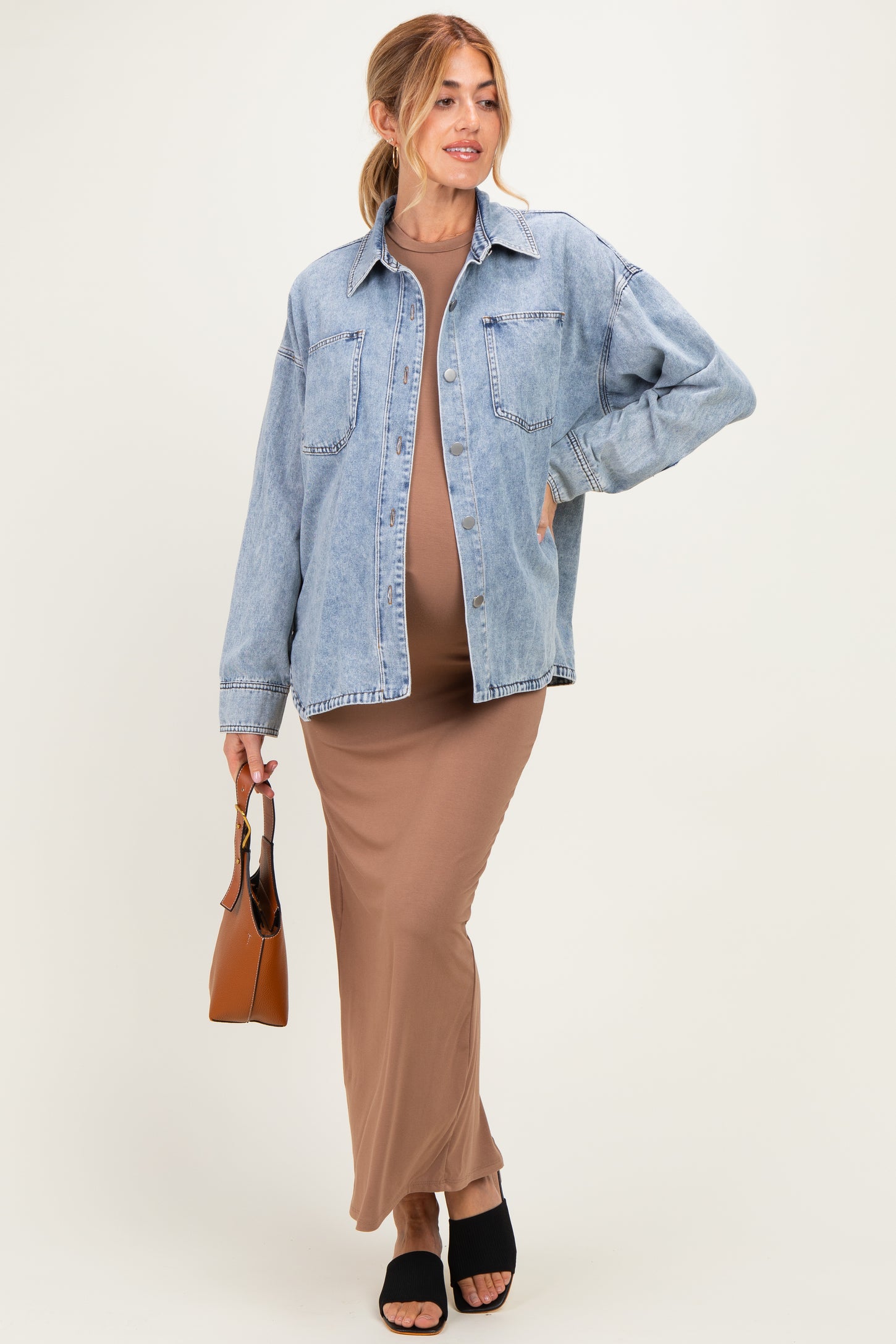 woman wearing a brown maternity maxi dress and a light blue oversized denim jacket, holding a brown handbag and showing her baby bump