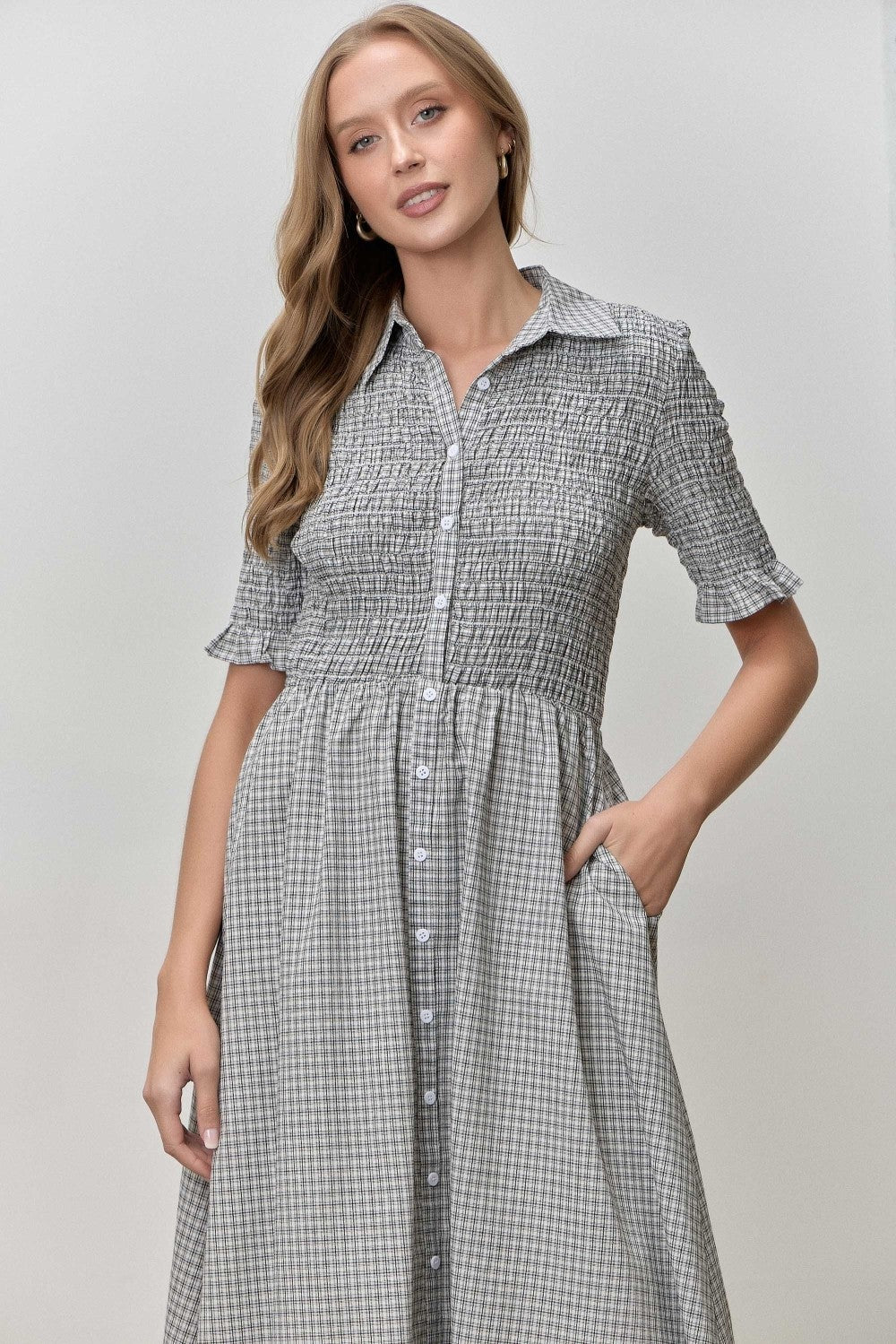 woman wearing a black and white gingham button-up dress with short sleeves and a smocked bodice, standing with one hand in her pocket