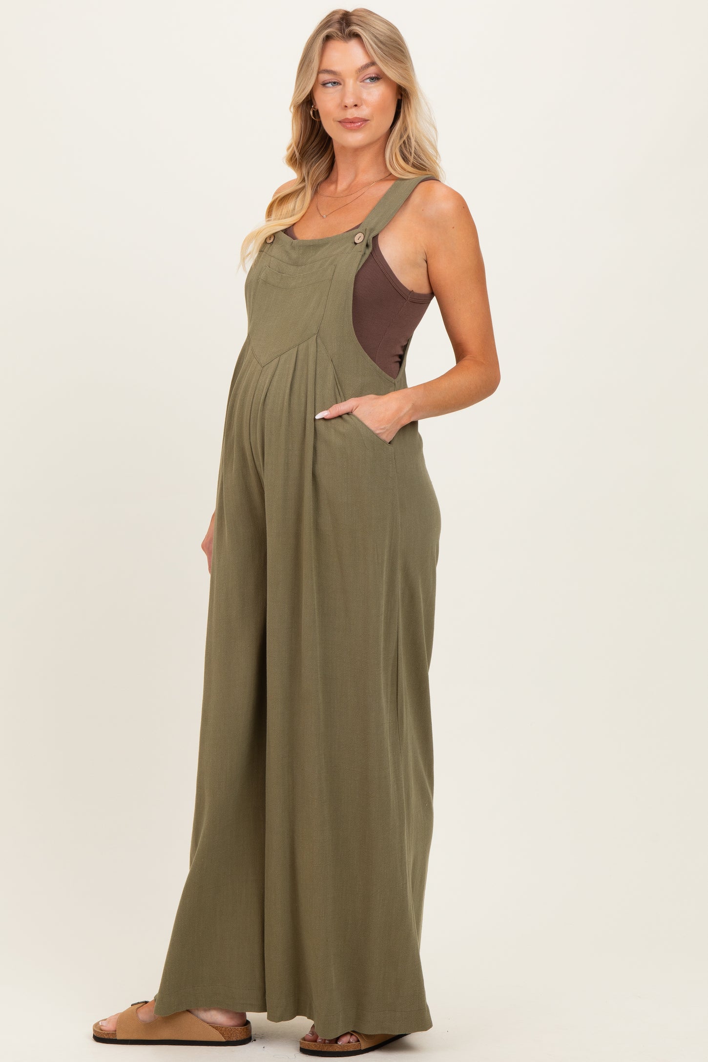 woman wearing an olive green maternity jumpsuit with pockets and a brown top underneath, showing her baby bump
