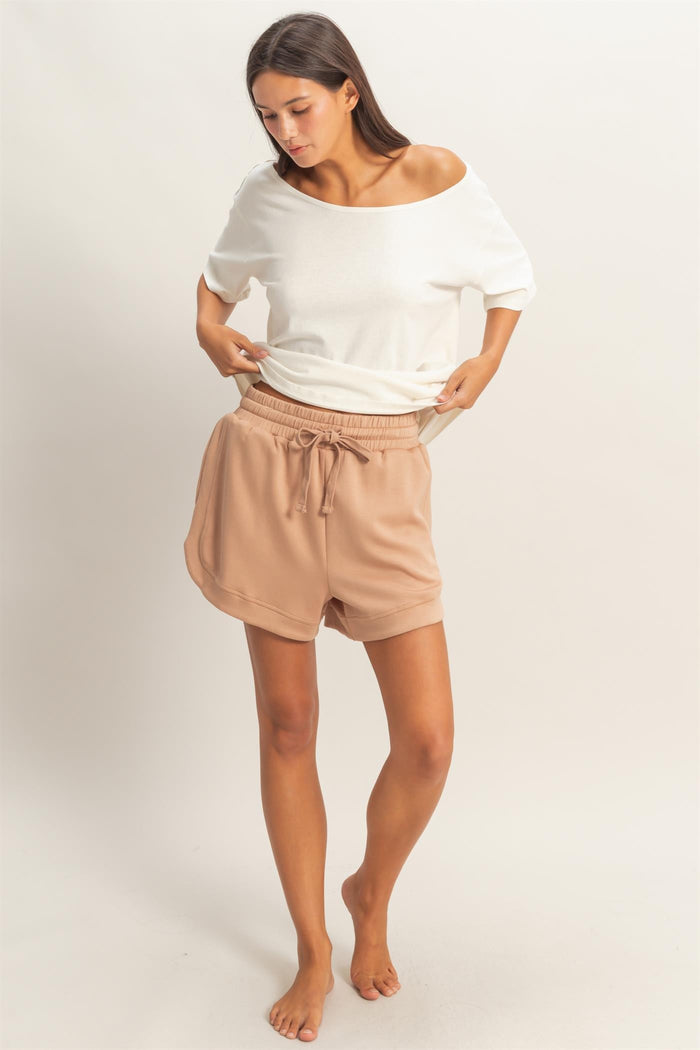 woman wearing a white off-shoulder top and beige drawstring shorts, standing barefoot and looking down