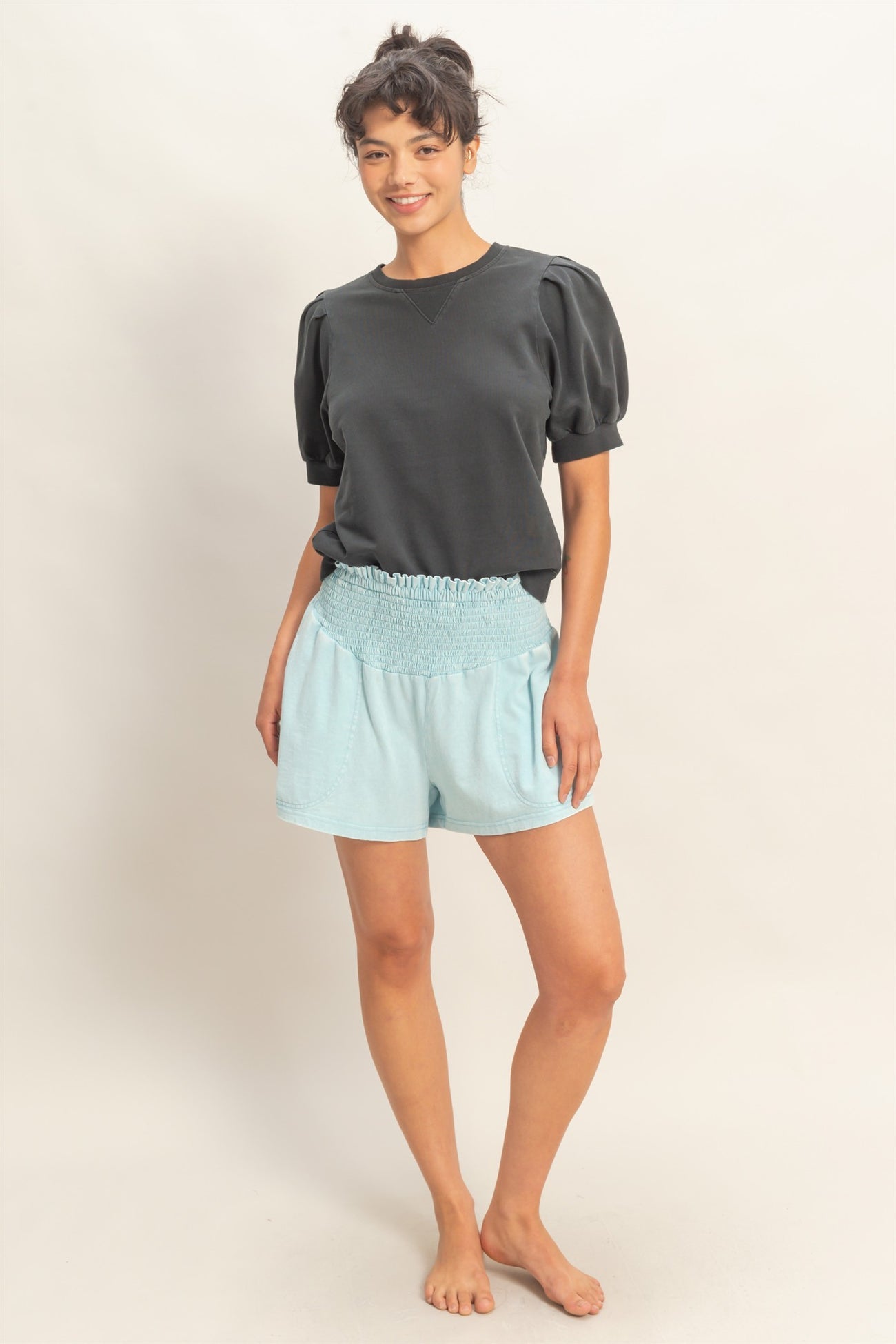 woman wearing a black puff sleeve sweatshirt and light blue smocked shorts, standing barefoot with a neutral background
