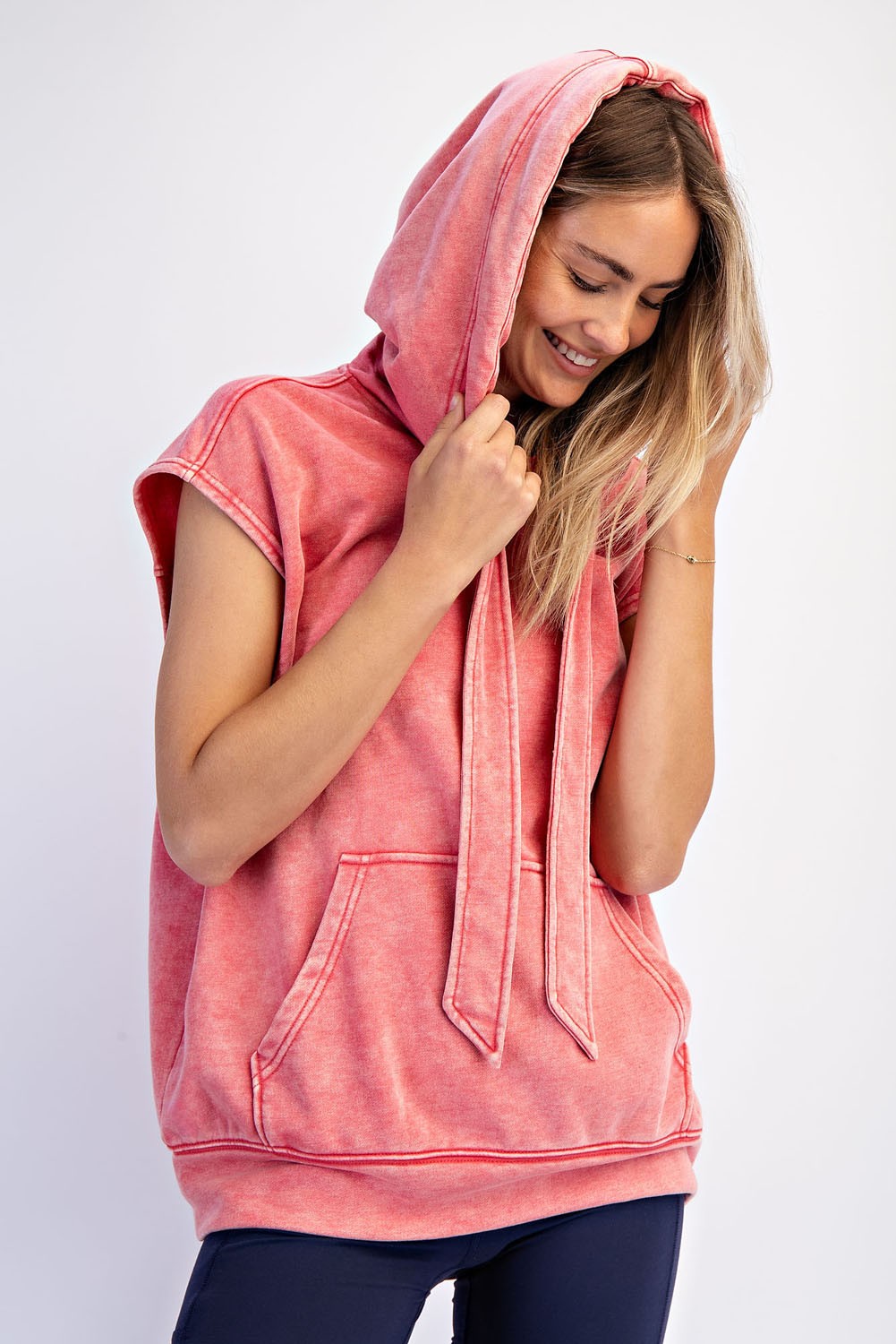 woman wearing a coral sleeveless oversized hoodie with a front pocket and tie detail on the hood
