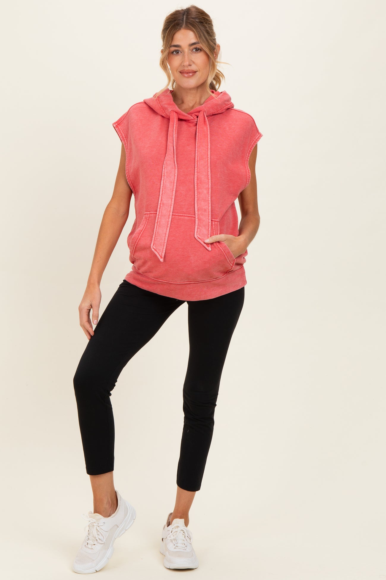 woman wearing a coral sleeveless maternity hoodie with a front pocket and black leggings