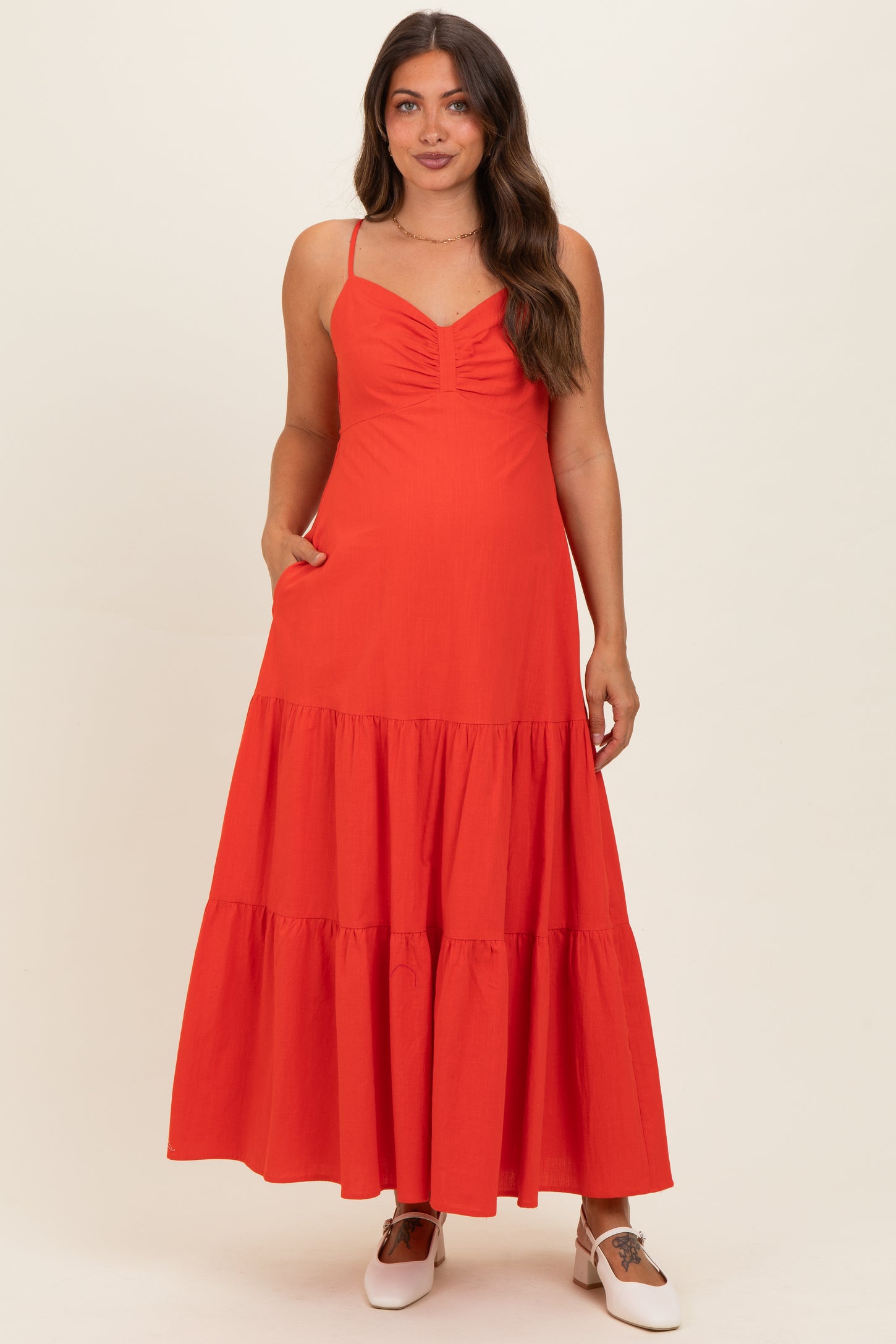 woman wearing a red maternity maxi dress with a ruched bodice and tiered skirt, standing with hands in pockets