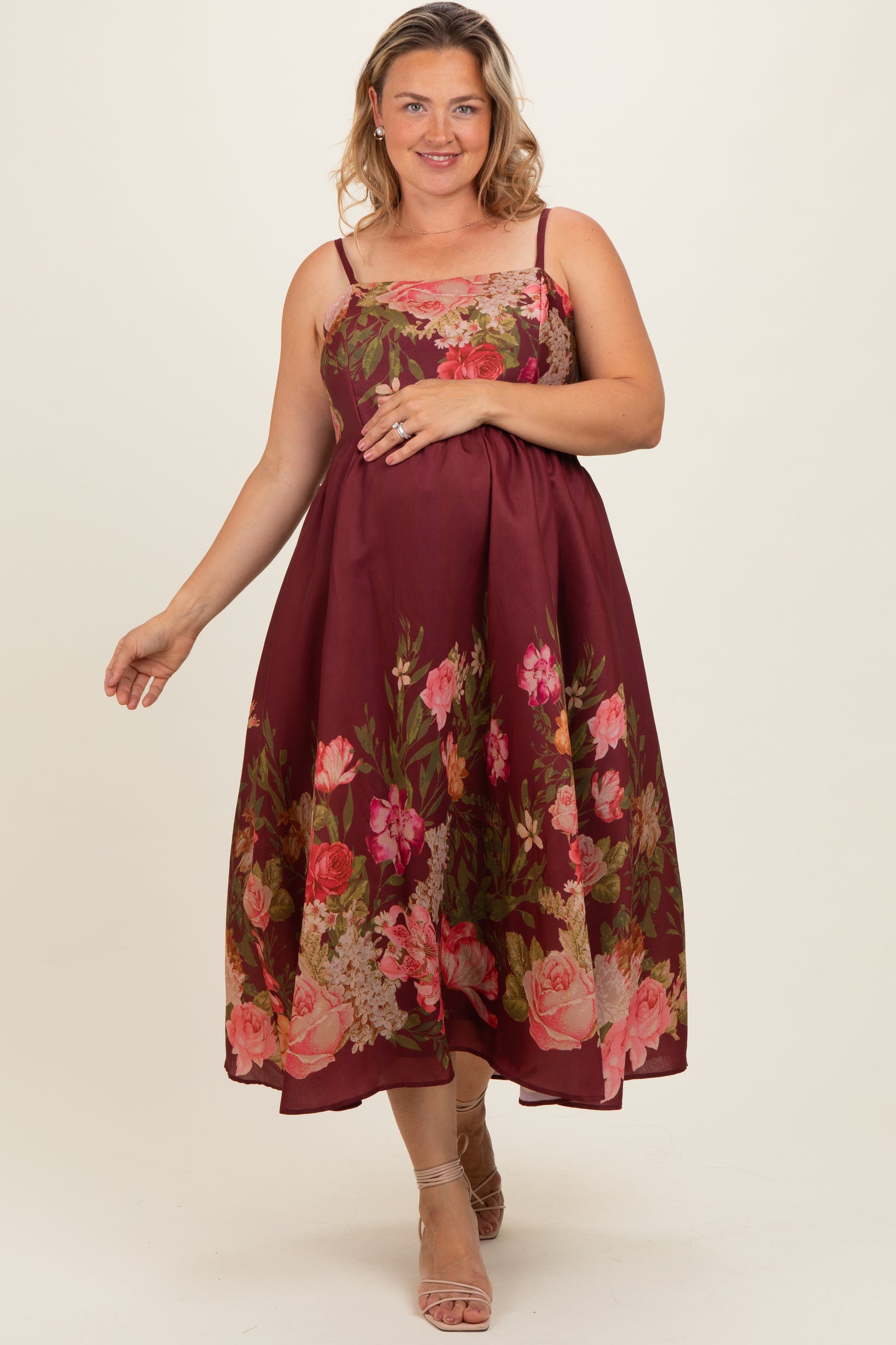 woman wearing a burgundy floral maternity plus dress with spaghetti straps and a flowing skirt, gently cradling her baby bump