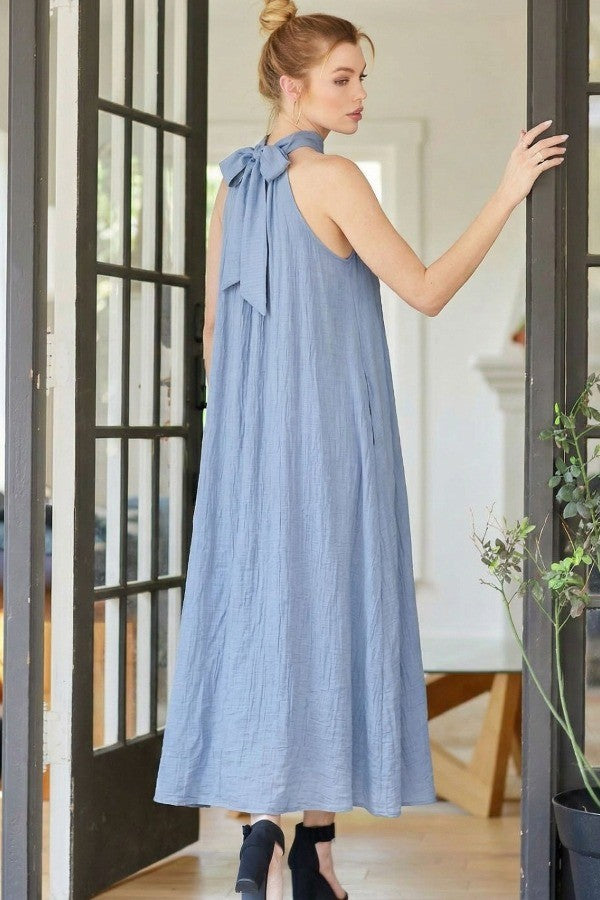 woman wearing a light blue sleeveless maxi dress with a bow detail at the back, standing by a door