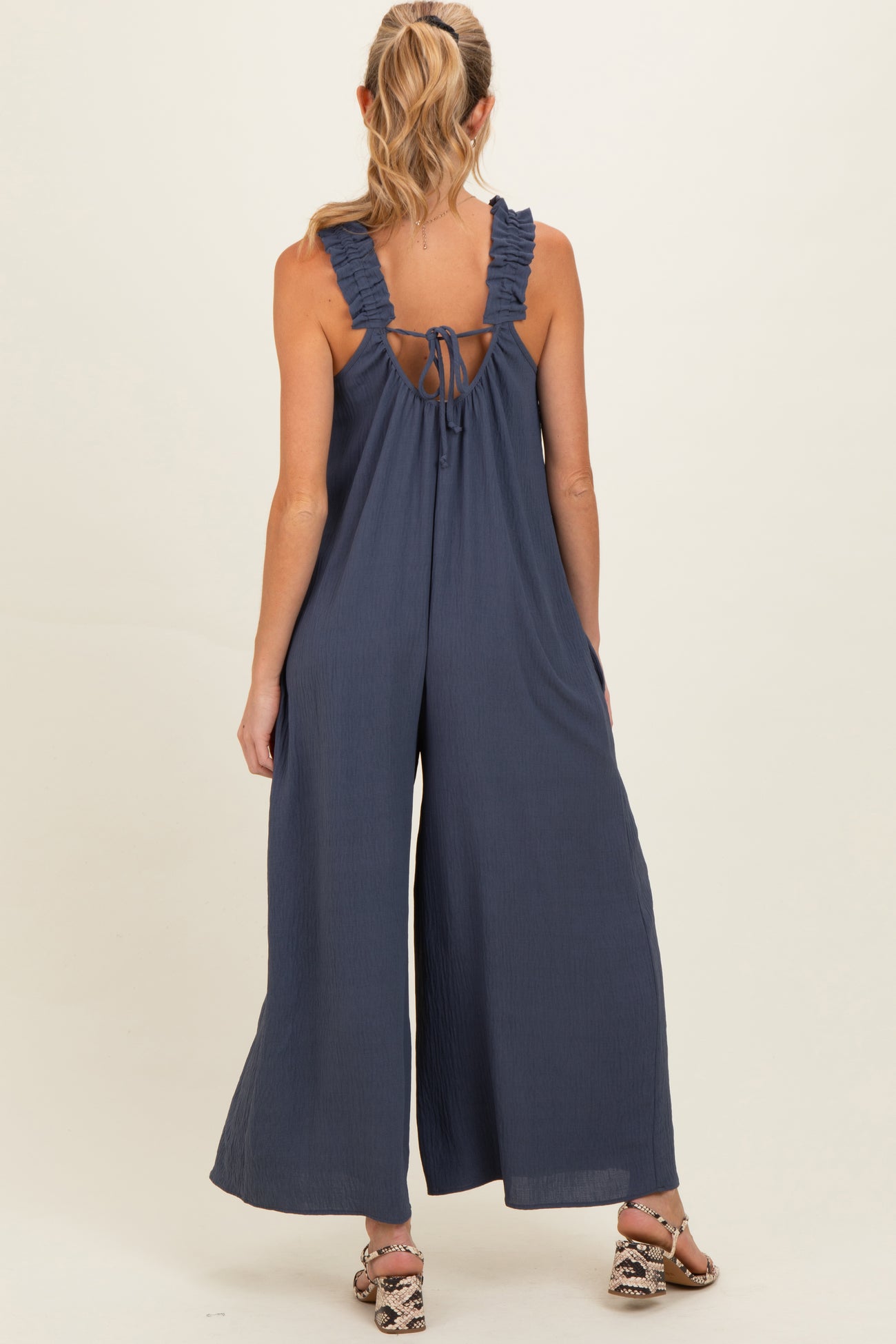 woman wearing a navy blue sleeveless maternity jumpsuit with ruffle straps and an open back design