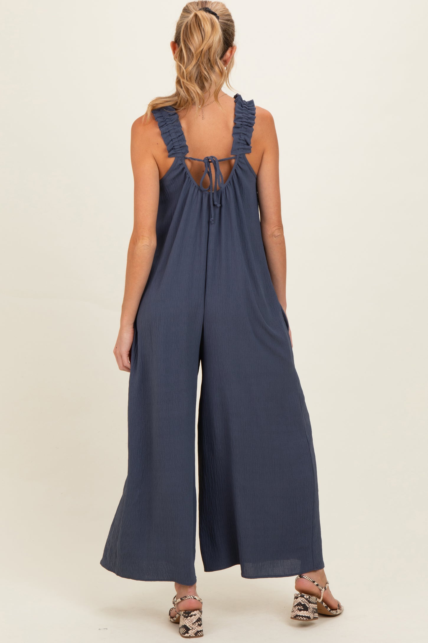 woman wearing a navy blue sleeveless maternity jumpsuit with ruffle straps and an open back design
