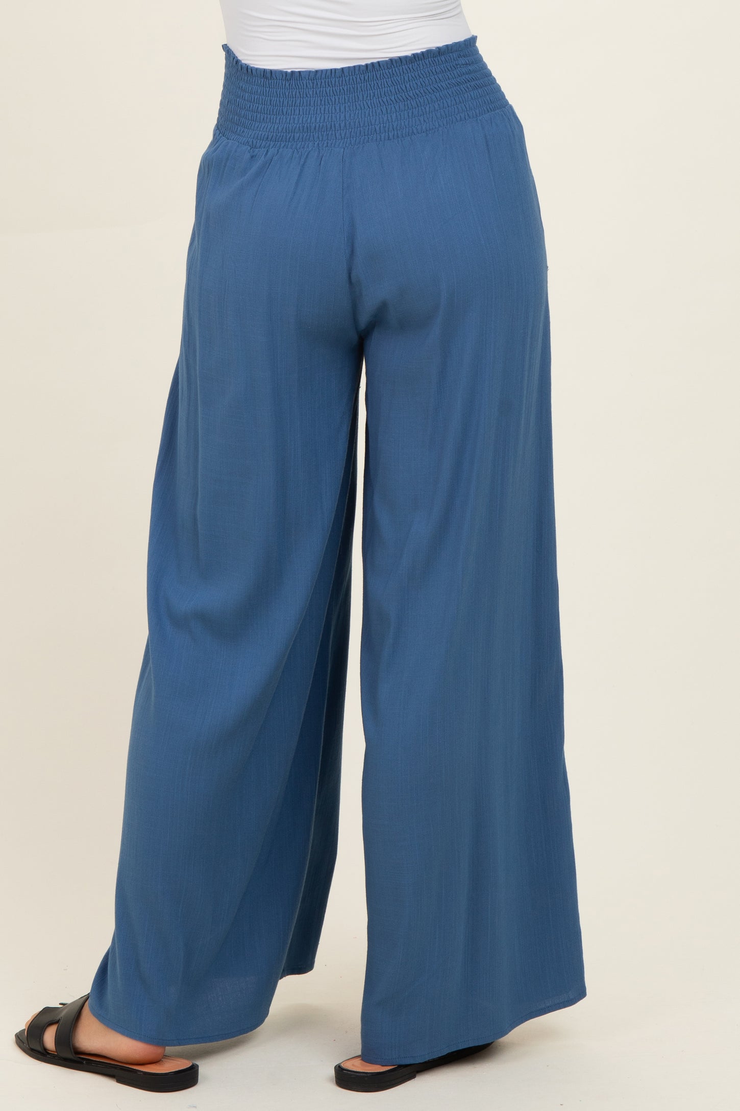 woman wearing a pair of blue wide-leg trousers with a smocked waistband and black sandals, viewed from the back