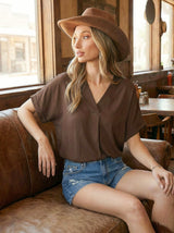 woman wearing a brown v-neck short sleeve top and denim shorts, sitting on a leather couch in a rustic setting