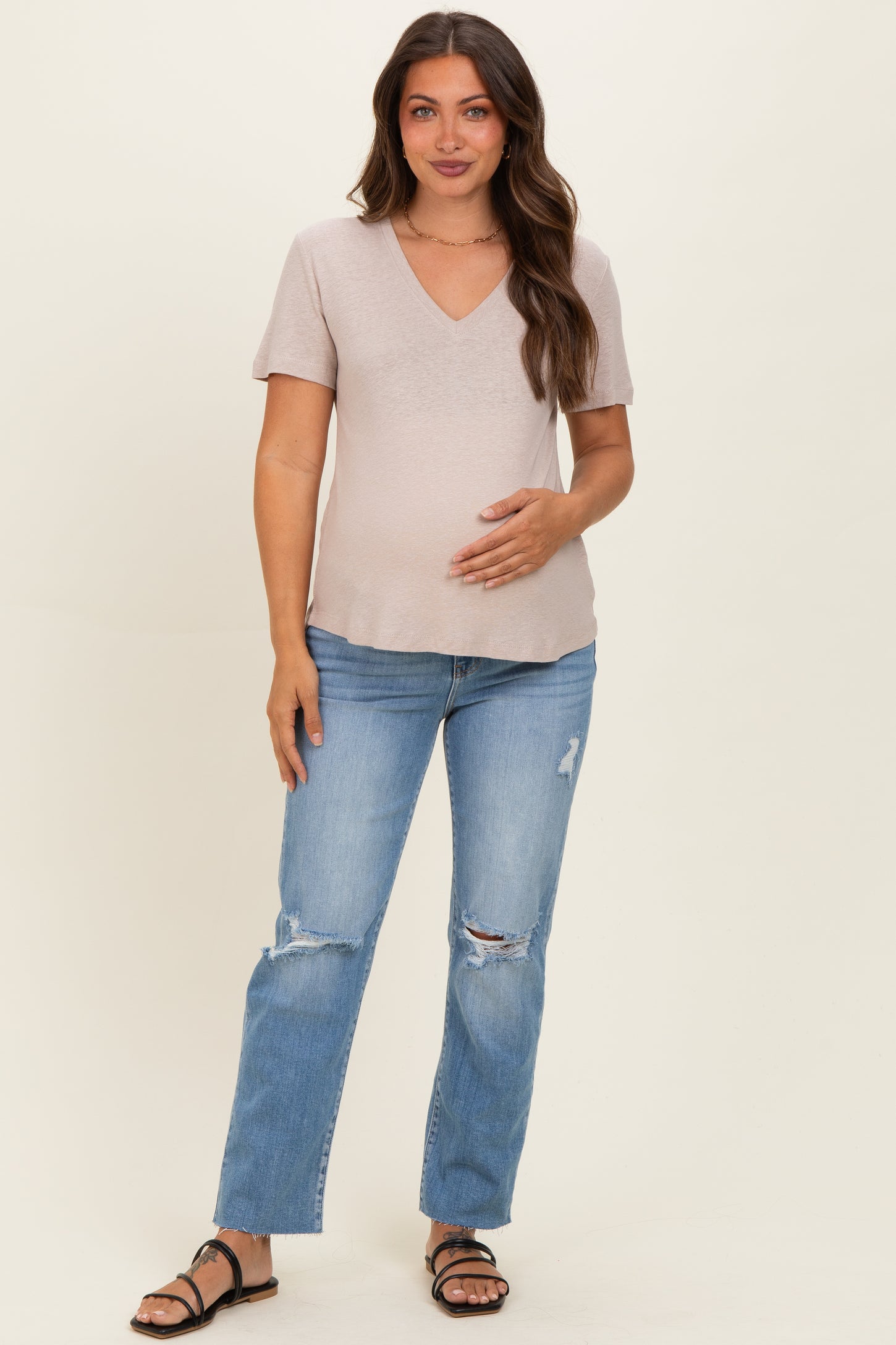 woman wearing a light pink short sleeve maternity top and distressed blue jeans, holding her baby bump
