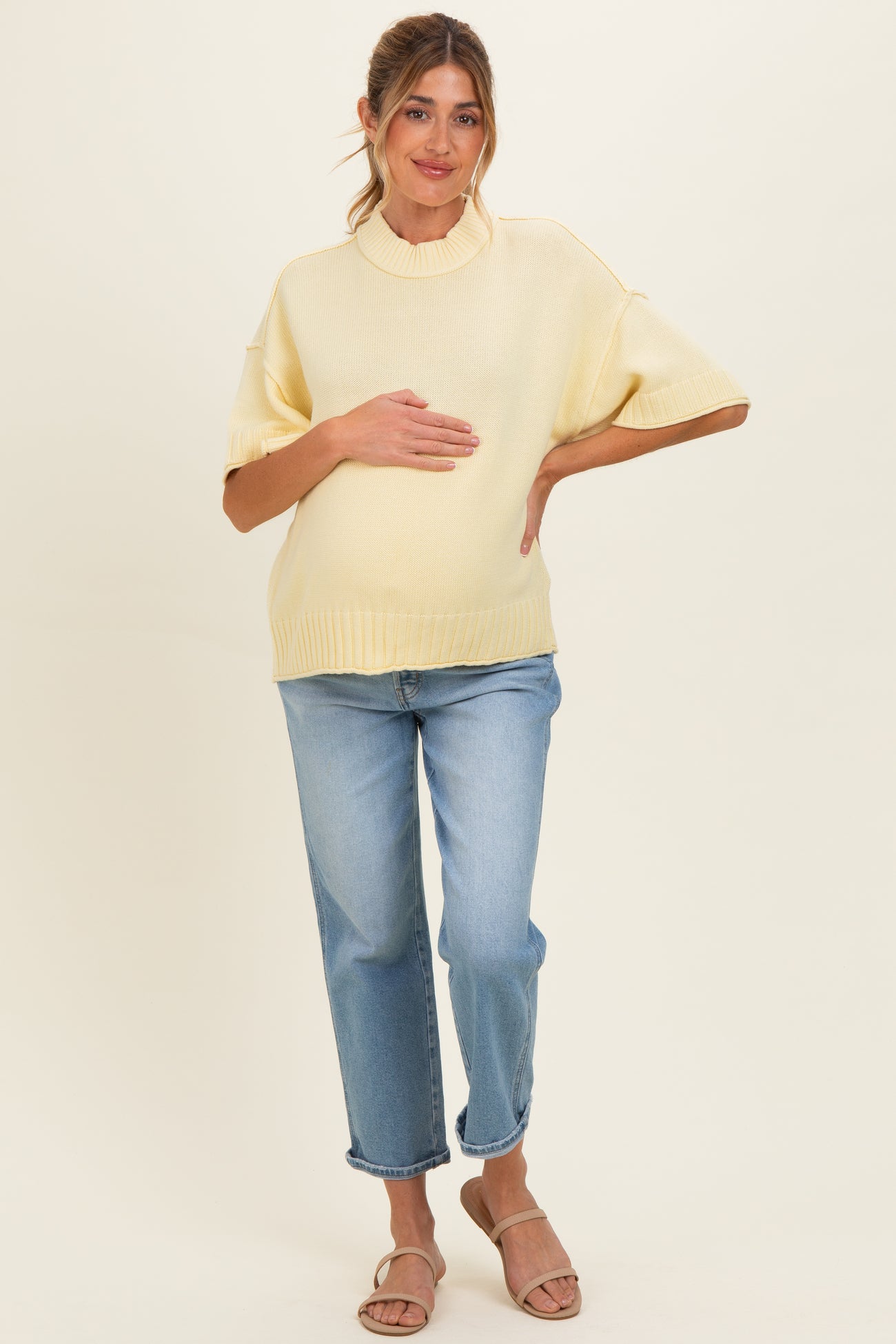 woman wearing a light yellow maternity sweater and blue jeans while holding her baby bump
