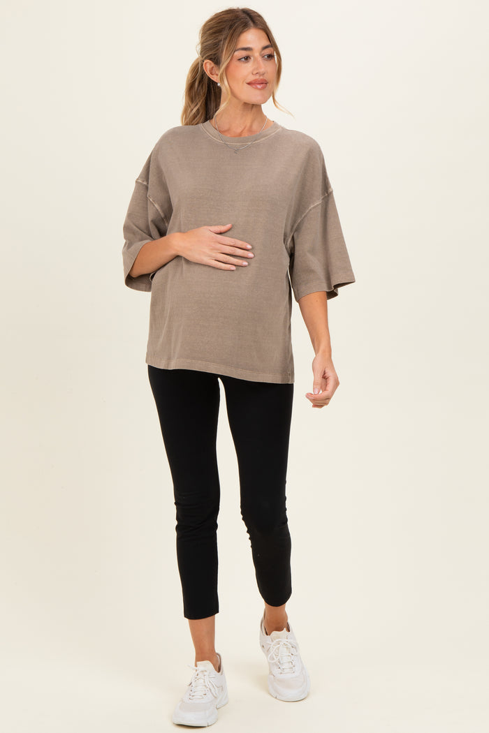 woman wearing a taupe oversized maternity top and black leggings, gently cradling her baby bump