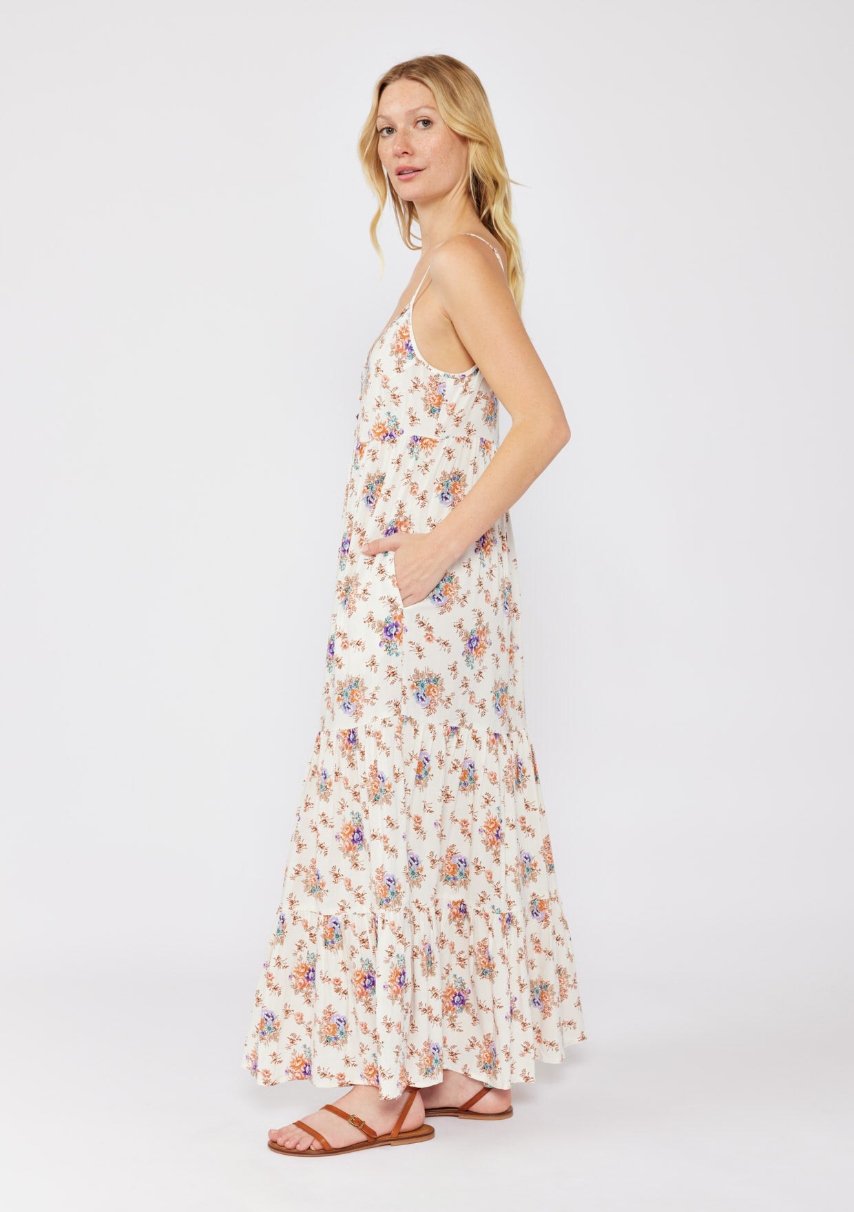 woman wearing a white floral maxi dress with thin straps and pockets, standing sideways with a light breeze in her hair
