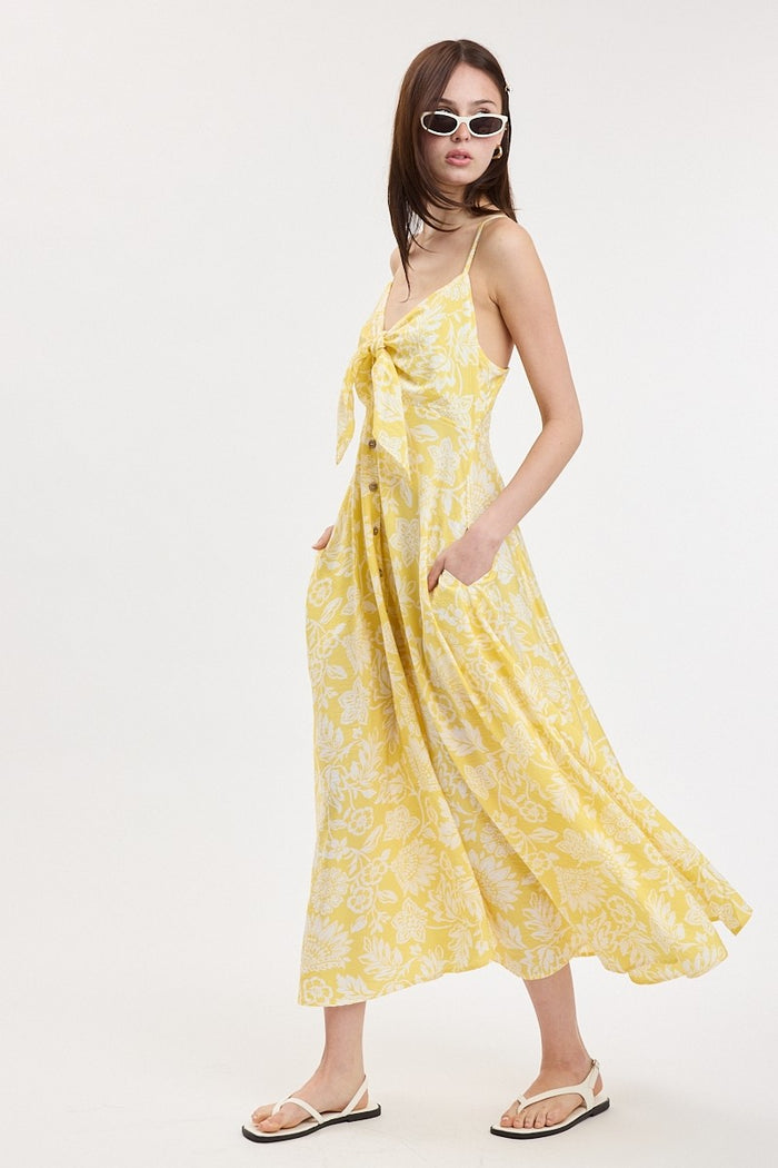 woman wearing a yellow floral print maxi dress with a tie front and side pockets, styled with white sunglasses and sandals