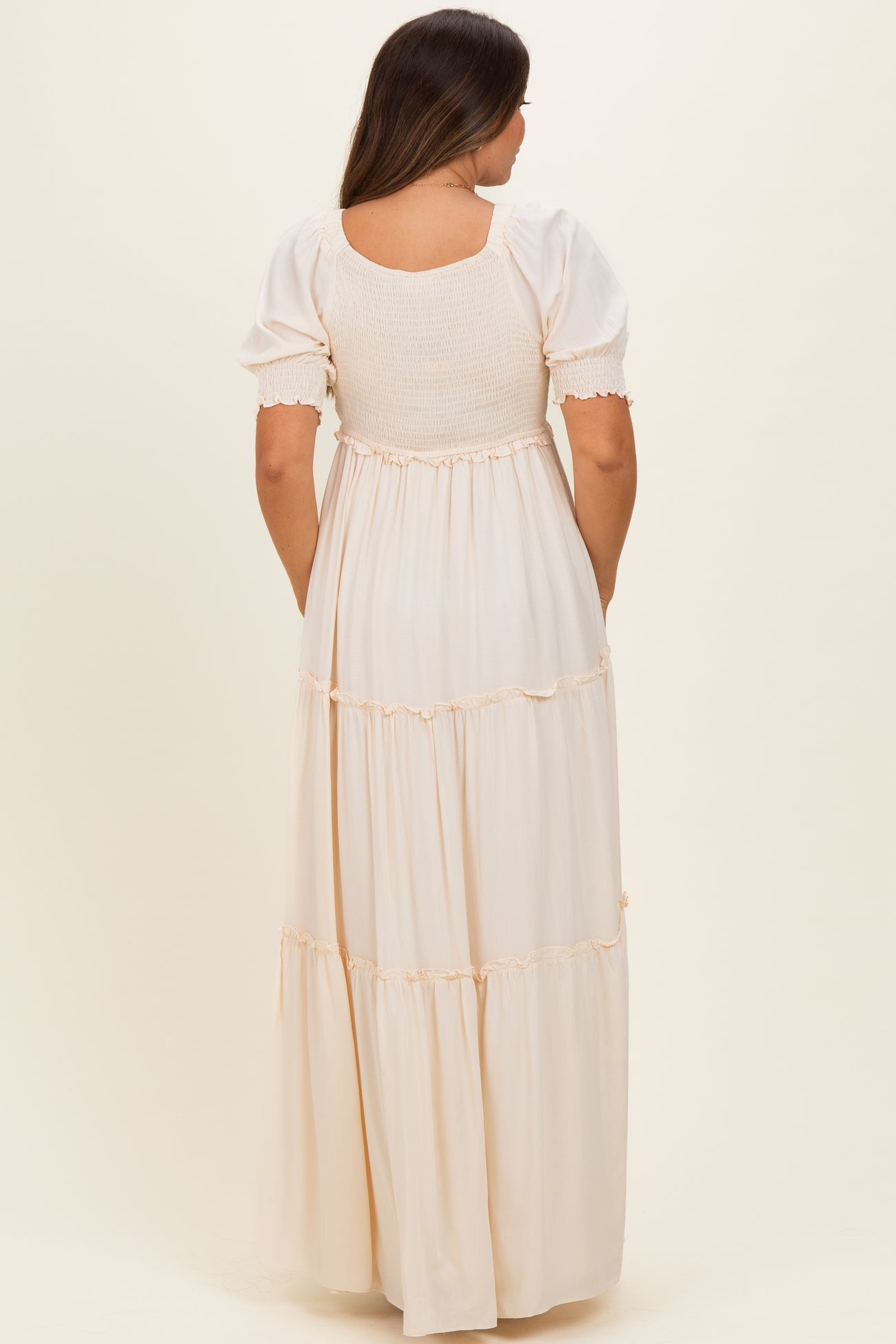 woman wearing a cream-colored maternity maxi dress with short puff sleeves and tiered skirt, showing a baby bump from the back