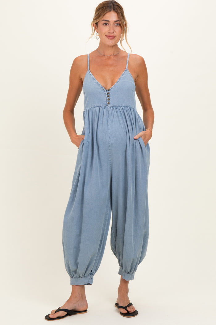 woman wearing a light blue maternity jumpsuit with a v-neckline, button details, and pockets, holding her baby bump