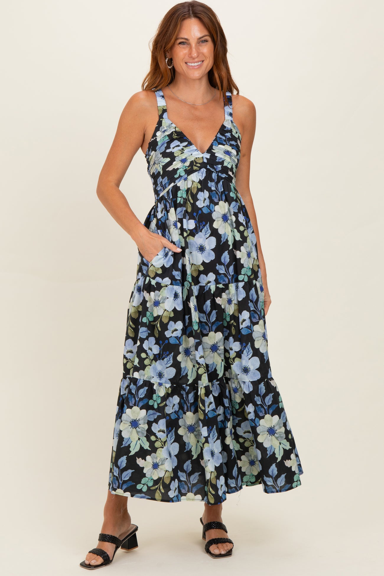 woman wearing a floral black maxi dress with blue and green flowers and pockets, posing with straps and sandals.