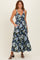 woman wearing a floral black maxi dress with blue and green flowers and pockets, posing with straps and sandals.