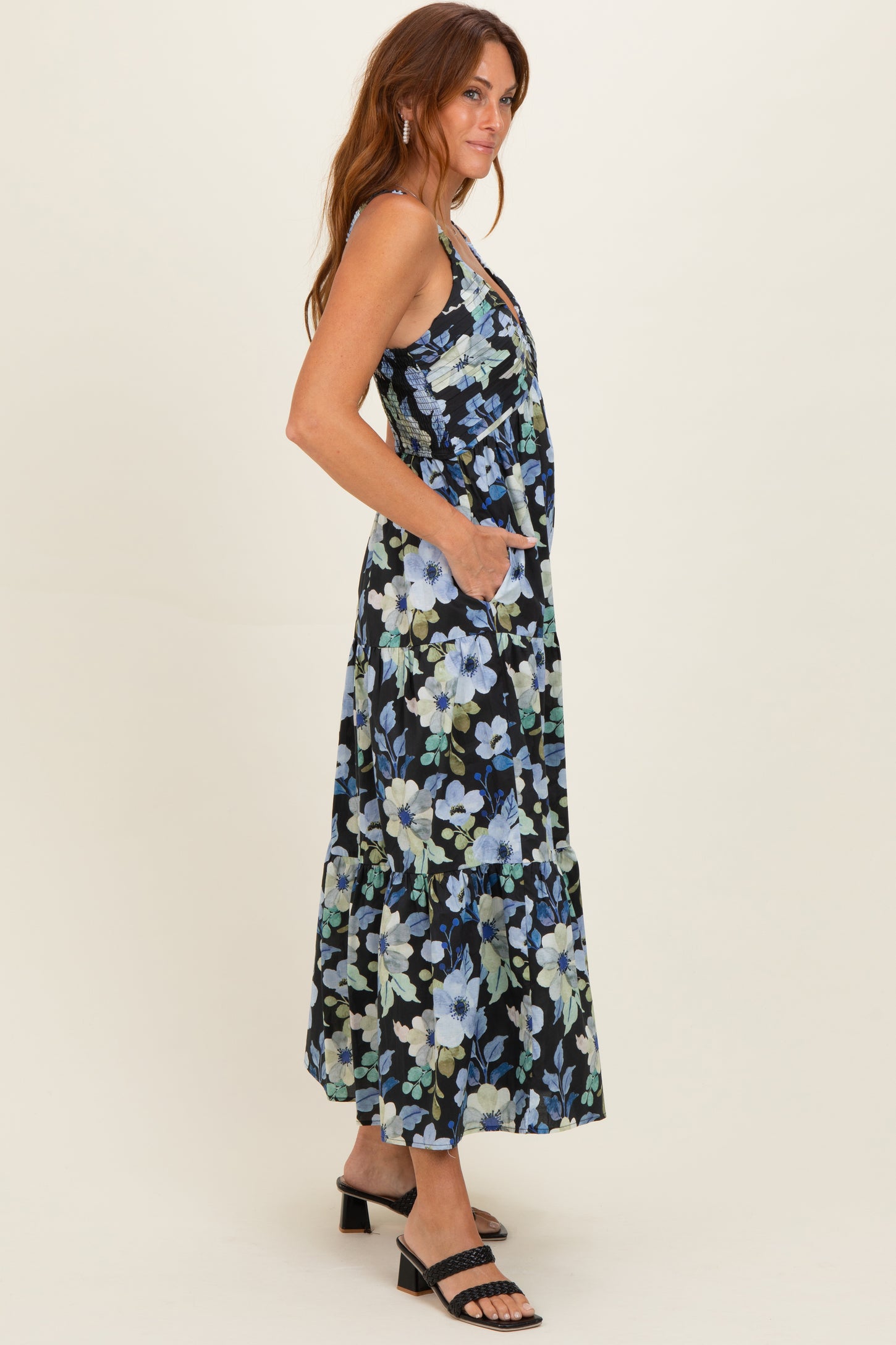 woman wearing a black floral maxi dress with pockets and a tiered skirt, standing sideways with a relaxed pose