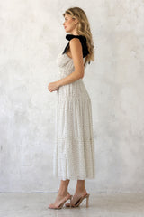 woman wearing a white polka dot midi dress with tiered skirt and black shoulder straps, standing sideways on a textured background