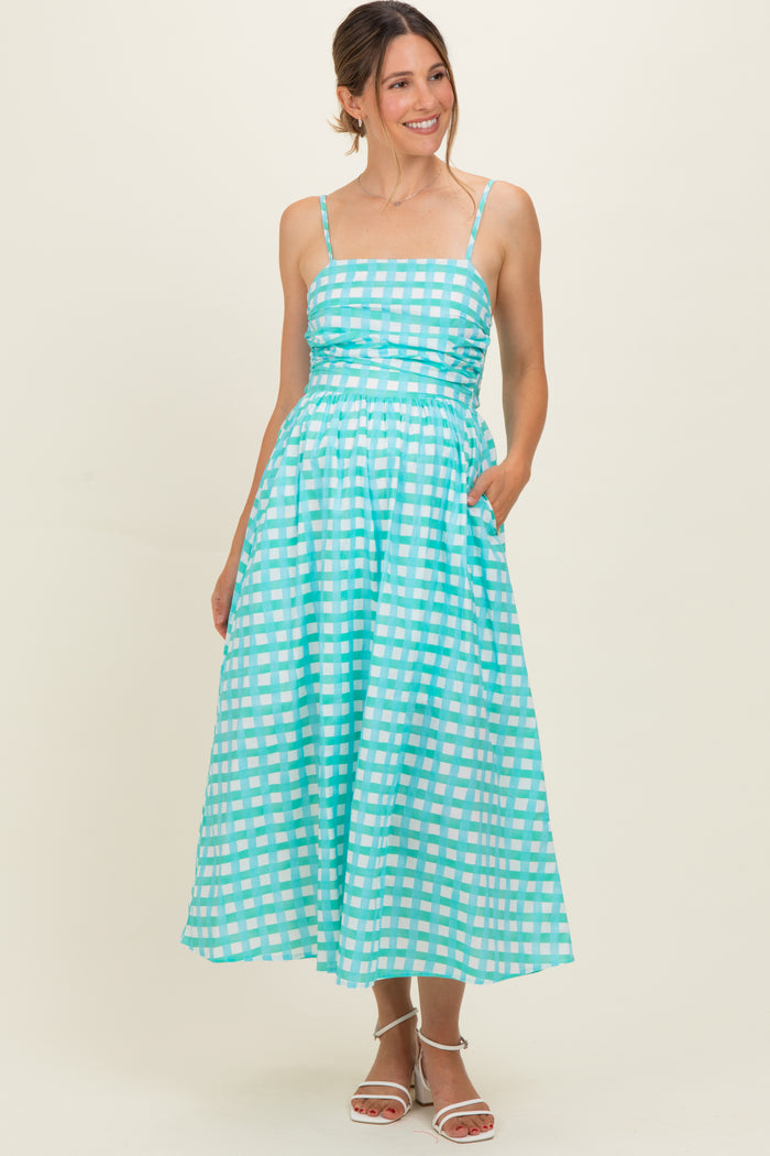 woman wearing a turquoise maternity dress with a checkered pattern and pockets, styled with spaghetti straps