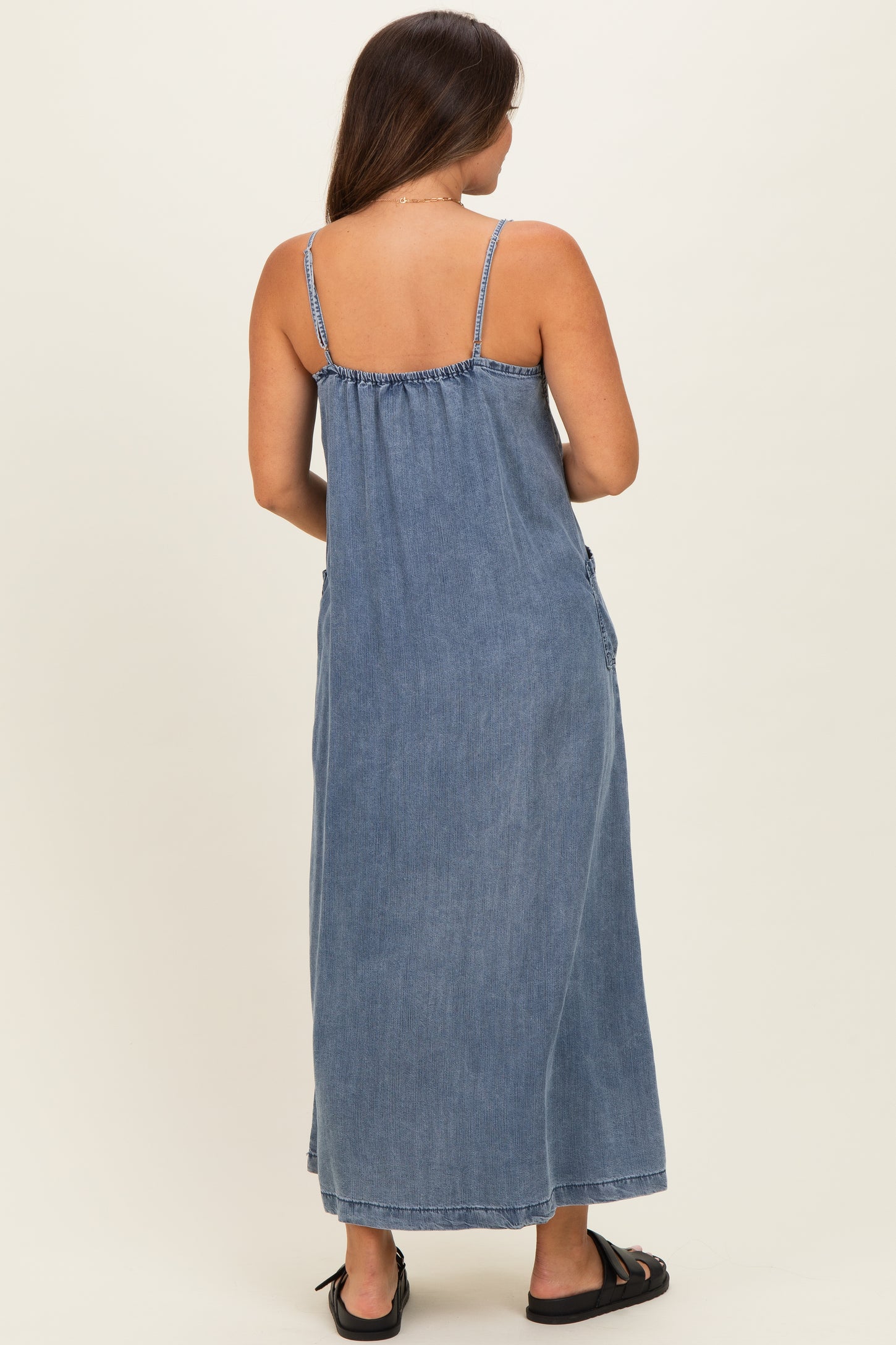 woman wearing a blue denim maxi maternity dress with adjustable straps, shown from the back