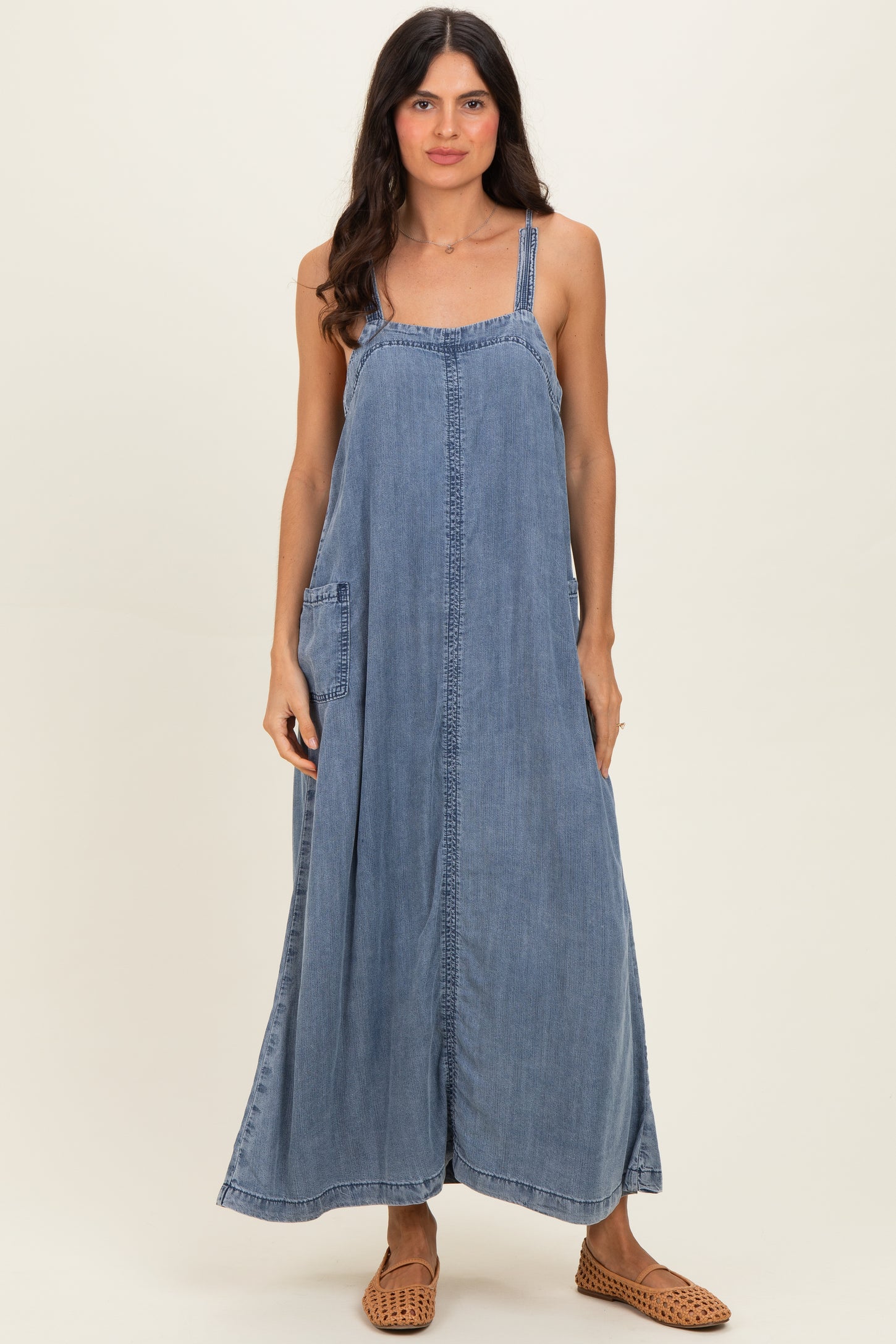 woman wearing a blue denim maxi dress with thin straps and side pockets, standing against a neutral background