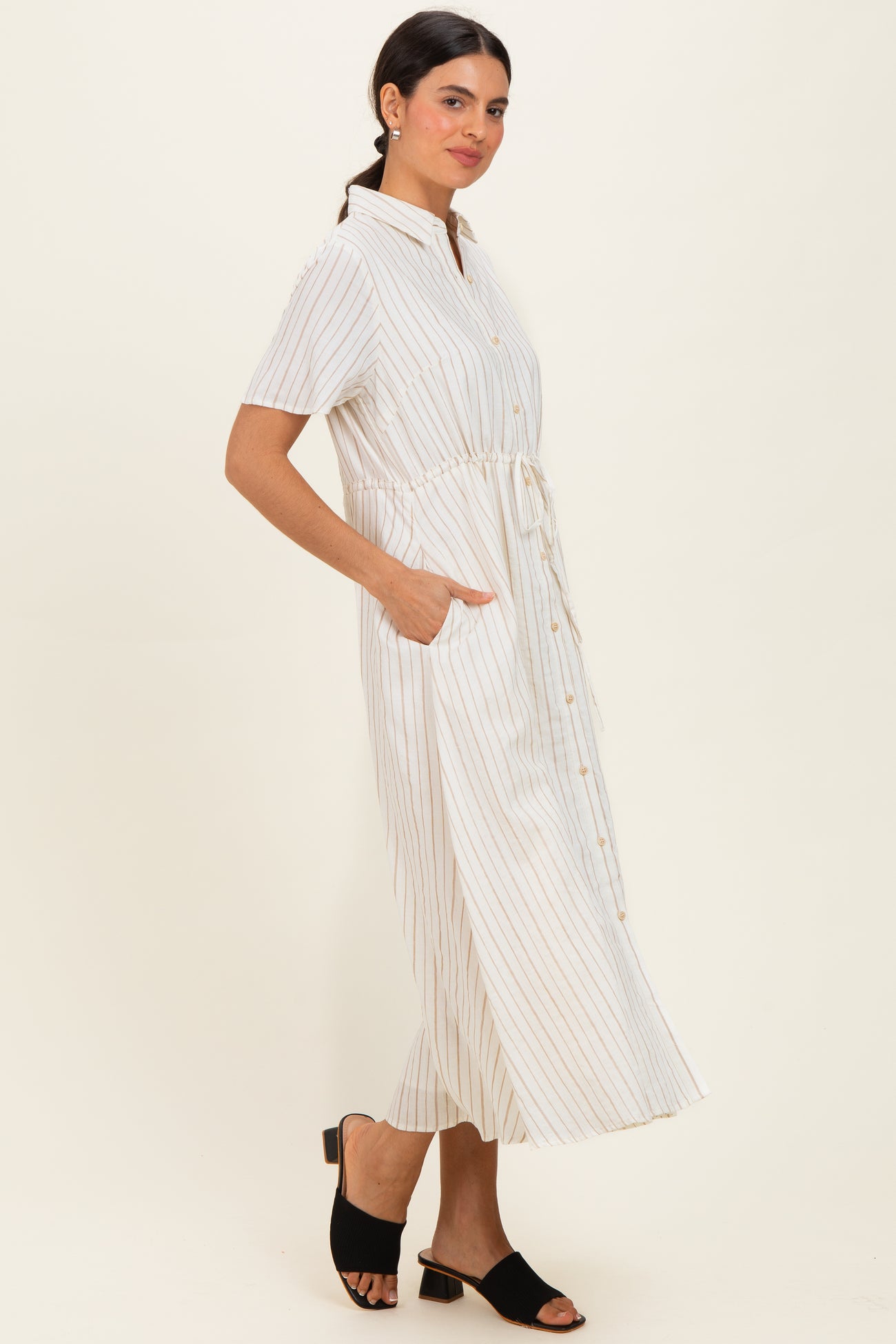 woman wearing a white and beige striped midi shirt dress with a tie waist and side pockets, standing in a relaxed pose