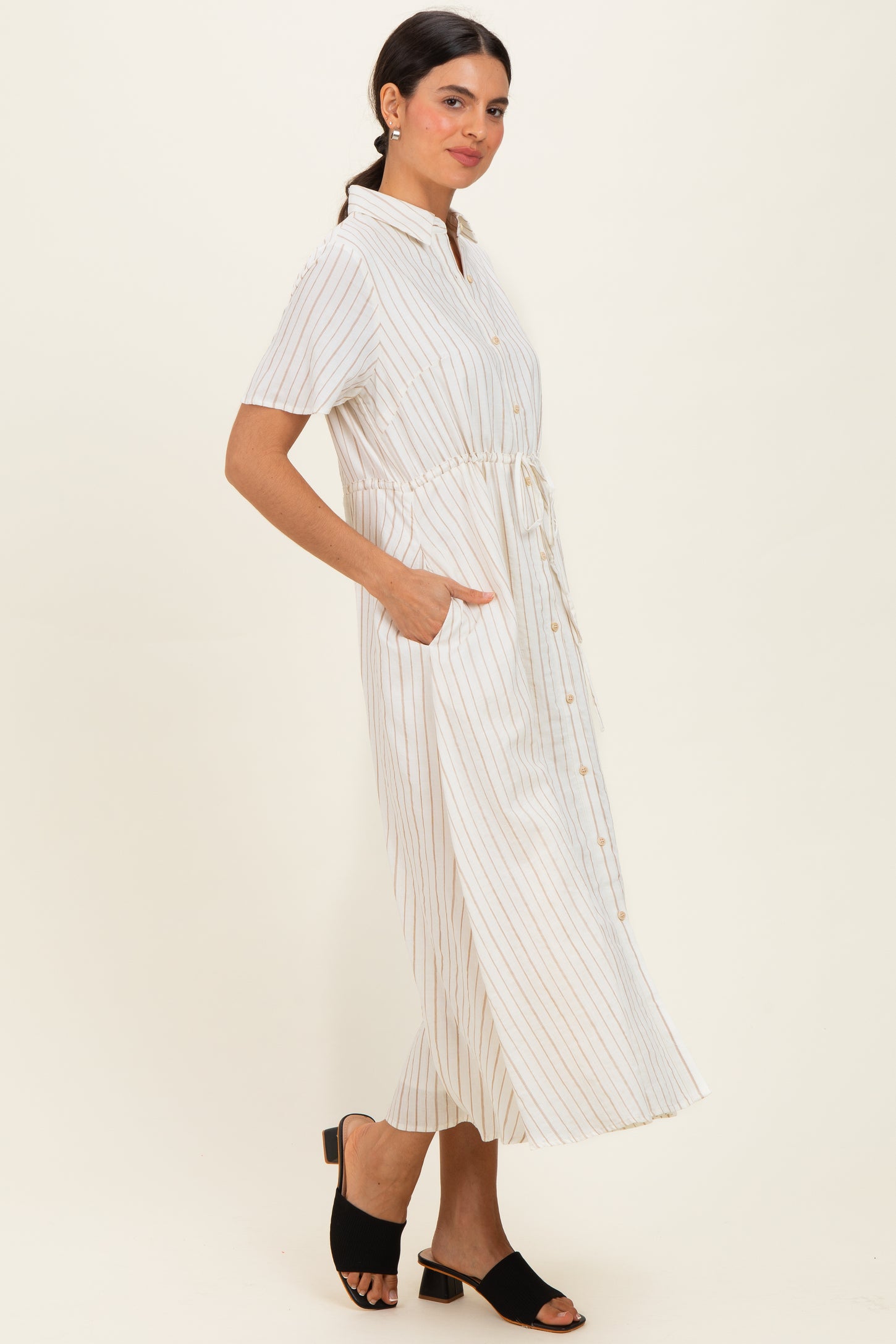woman wearing a white and beige striped midi shirt dress with a tie waist and side pockets, standing in a relaxed pose