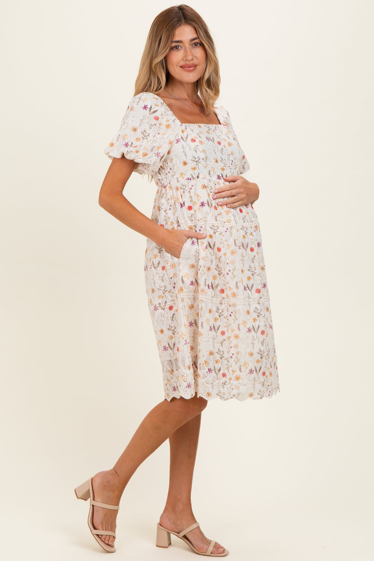 woman wearing a white floral maternity dress with short puff sleeves and pockets, gently cradling her baby bump