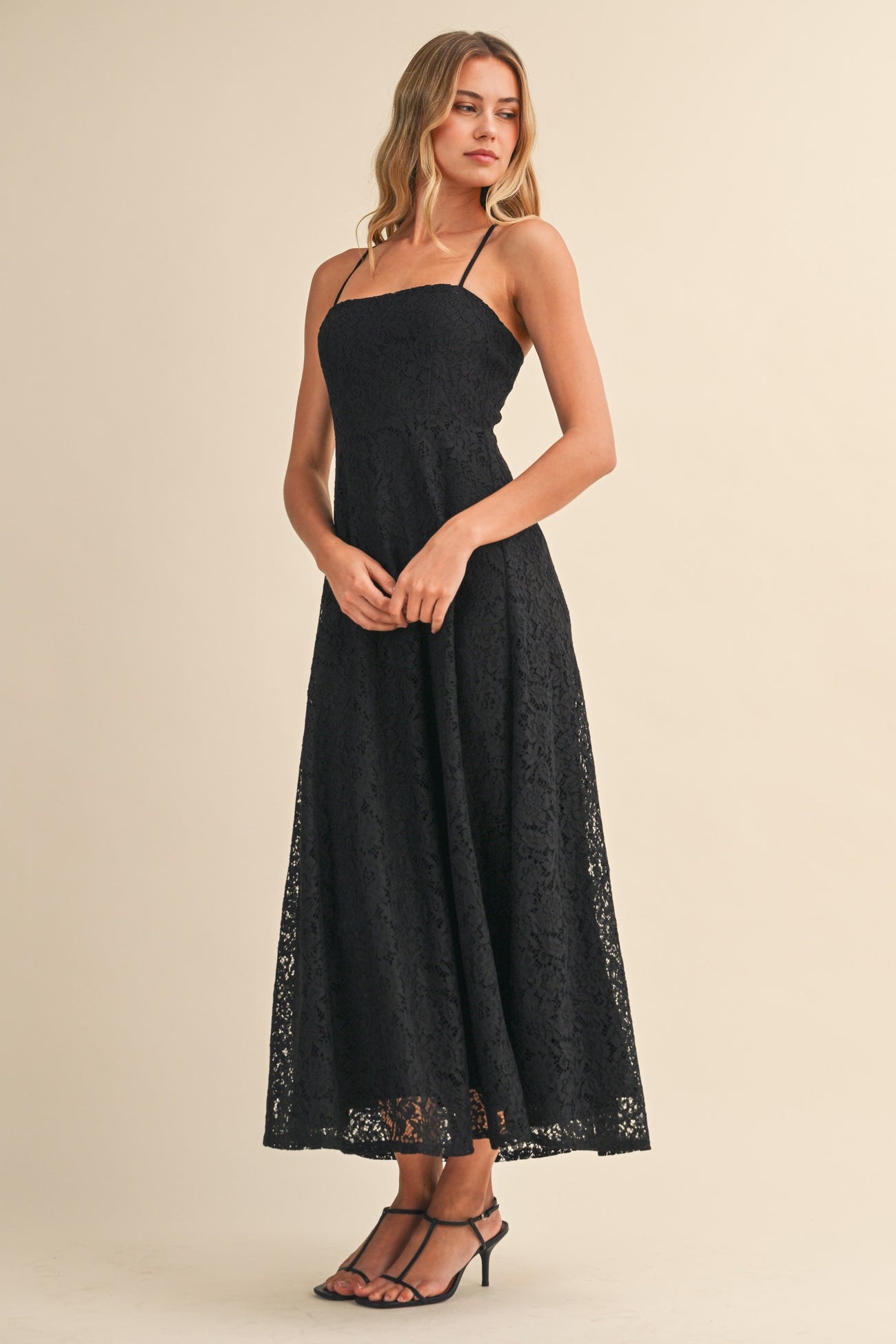 woman wearing a black lace maxi dress with spaghetti straps and a flowy skirt, standing elegantly with hands clasped