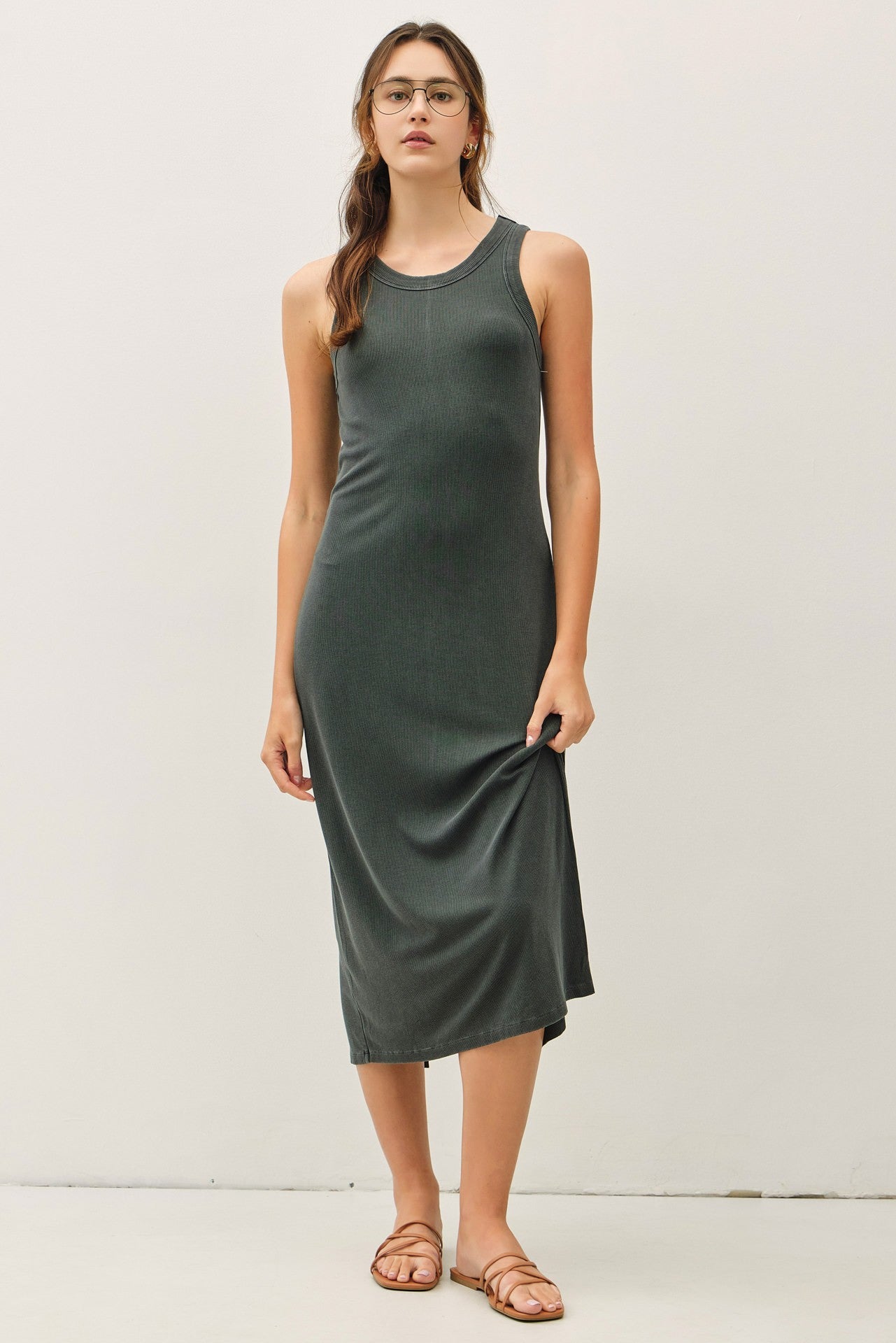 woman wearing a dark green sleeveless midi dress and holding the hem while standing against a plain background