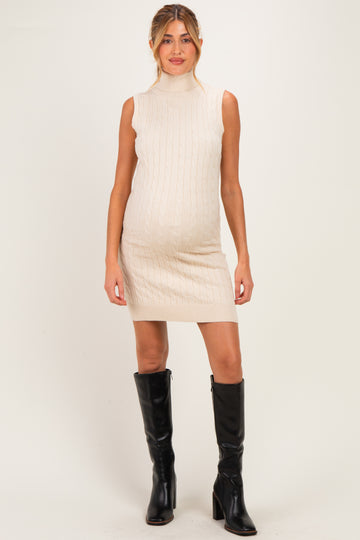 woman wearing a beige sleeveless maternity dress with cable knit pattern and a visible baby bump, paired with black knee-high boots