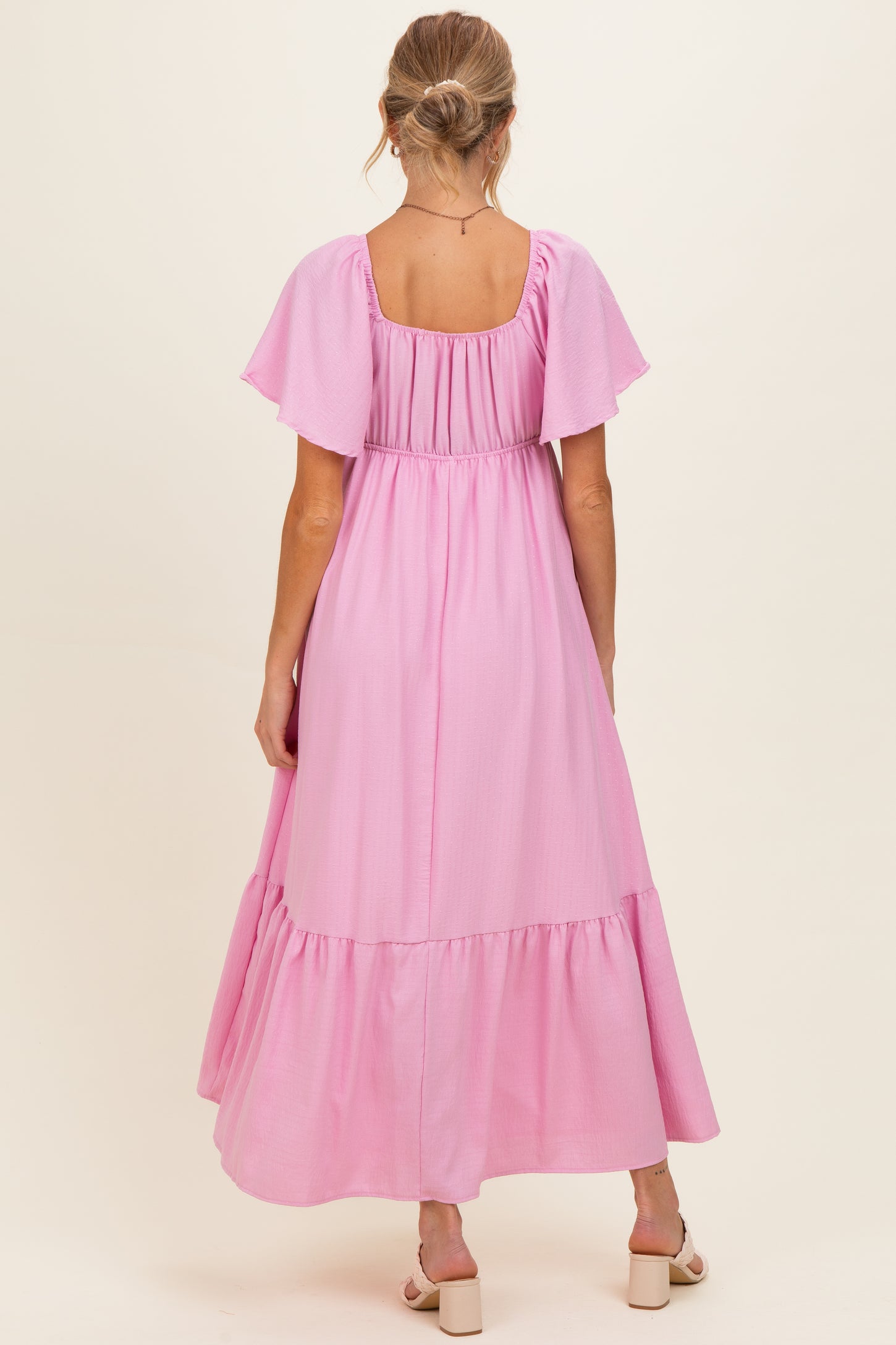 woman wearing a light pink maternity maxi dress with flutter sleeves and a tiered skirt, showing her baby bump from the back