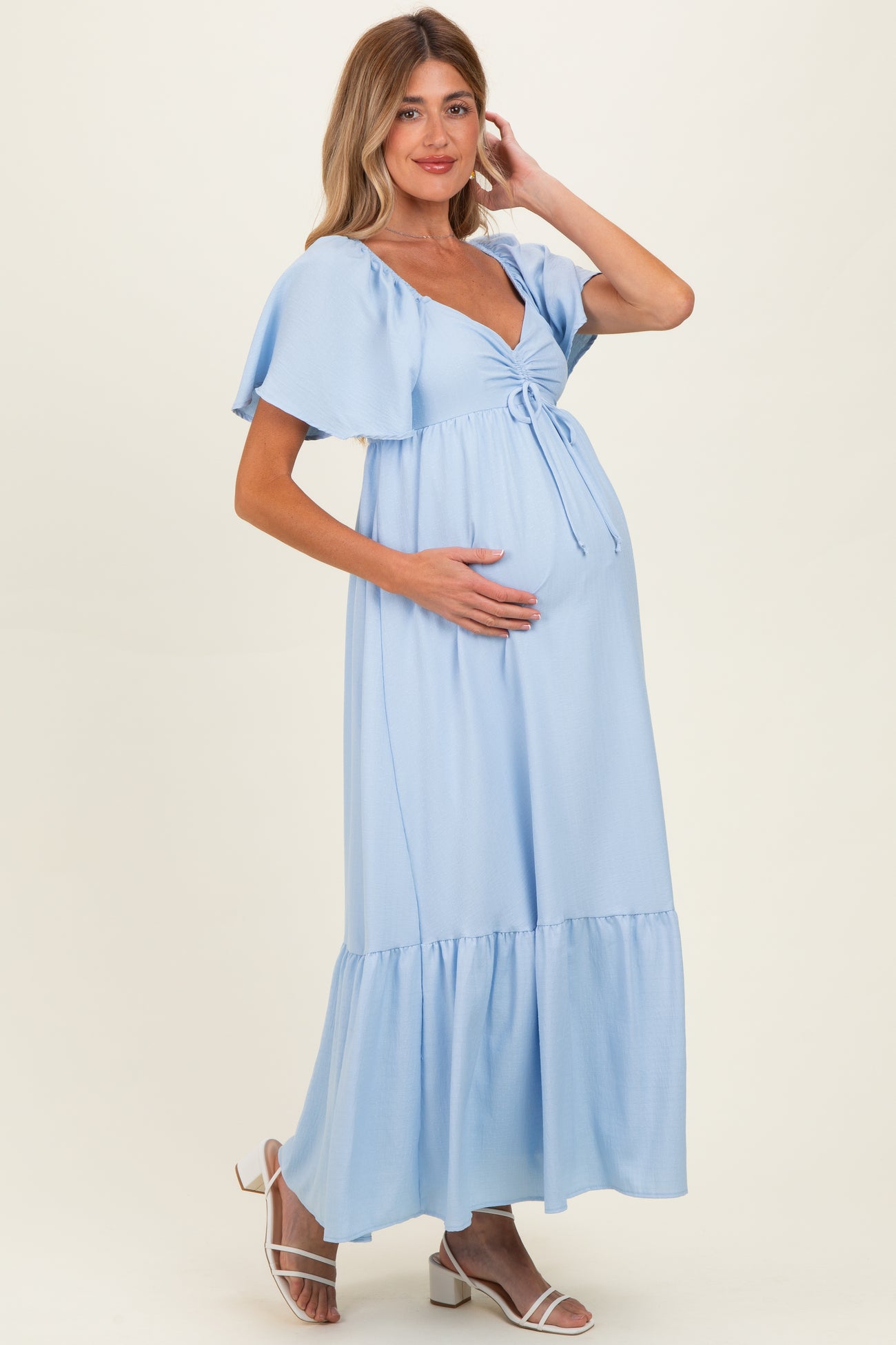 woman wearing a light blue maternity maxi dress with ruffled sleeves and a tie front, cradling her baby bump