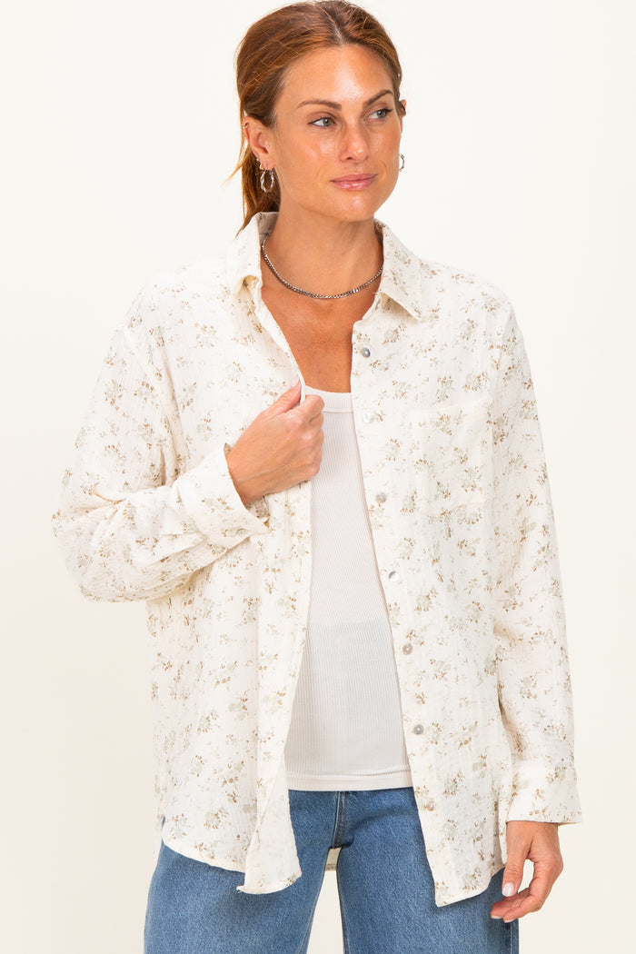 woman wearing a cream floral button-up shirt over a white tank top and blue jeans