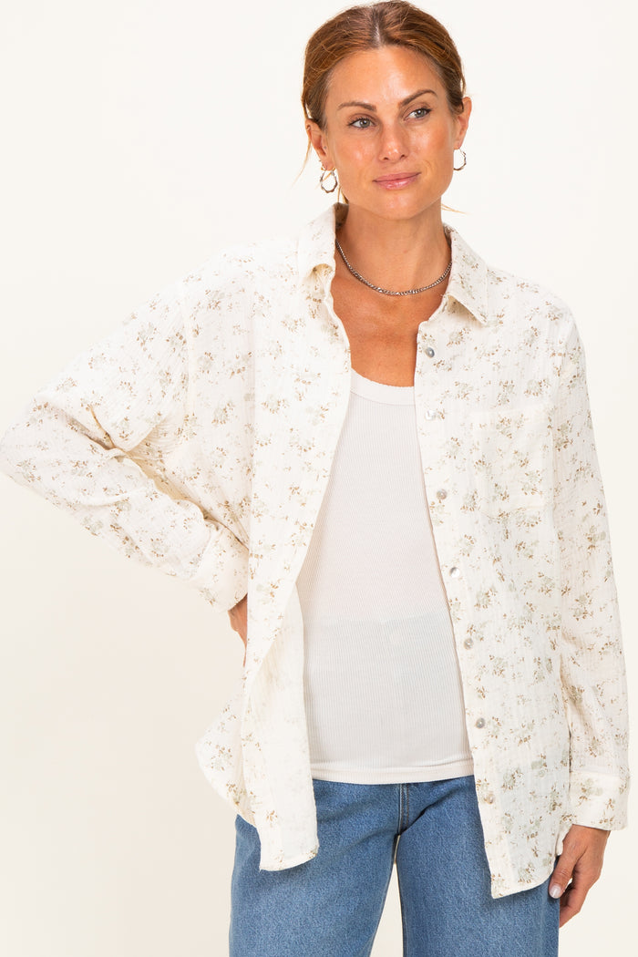 woman wearing a cream floral button-up shirt over a white tank top and blue jeans