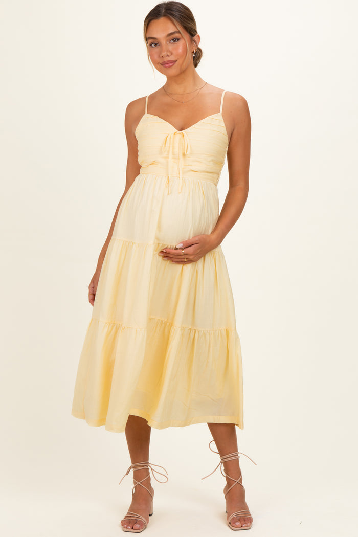 woman wearing a light yellow maternity dress with tie front and tiered skirt, holding her baby bump