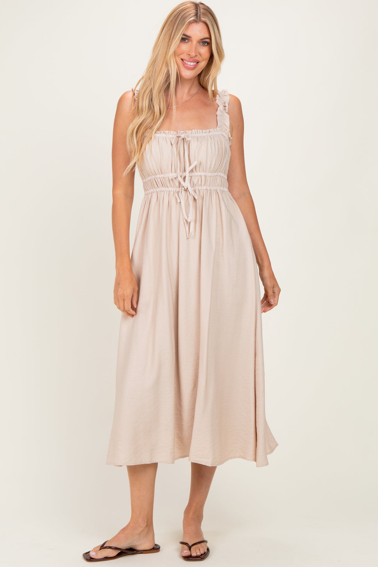 woman wearing a light beige maxi dress with adjustable straps and a drawstring waist, standing and smiling