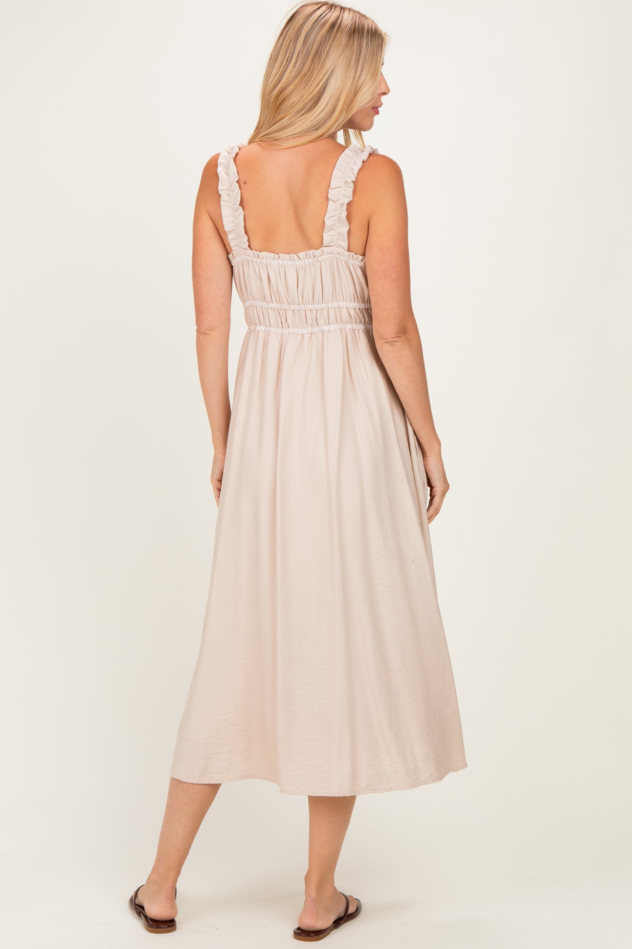 woman wearing a light beige midi dress with ruffle straps and a smocked back, standing with her back to the camera