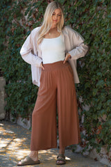 woman wearing a white tank top, oversized striped shirt, and brown wide leg pants standing in front of greenery