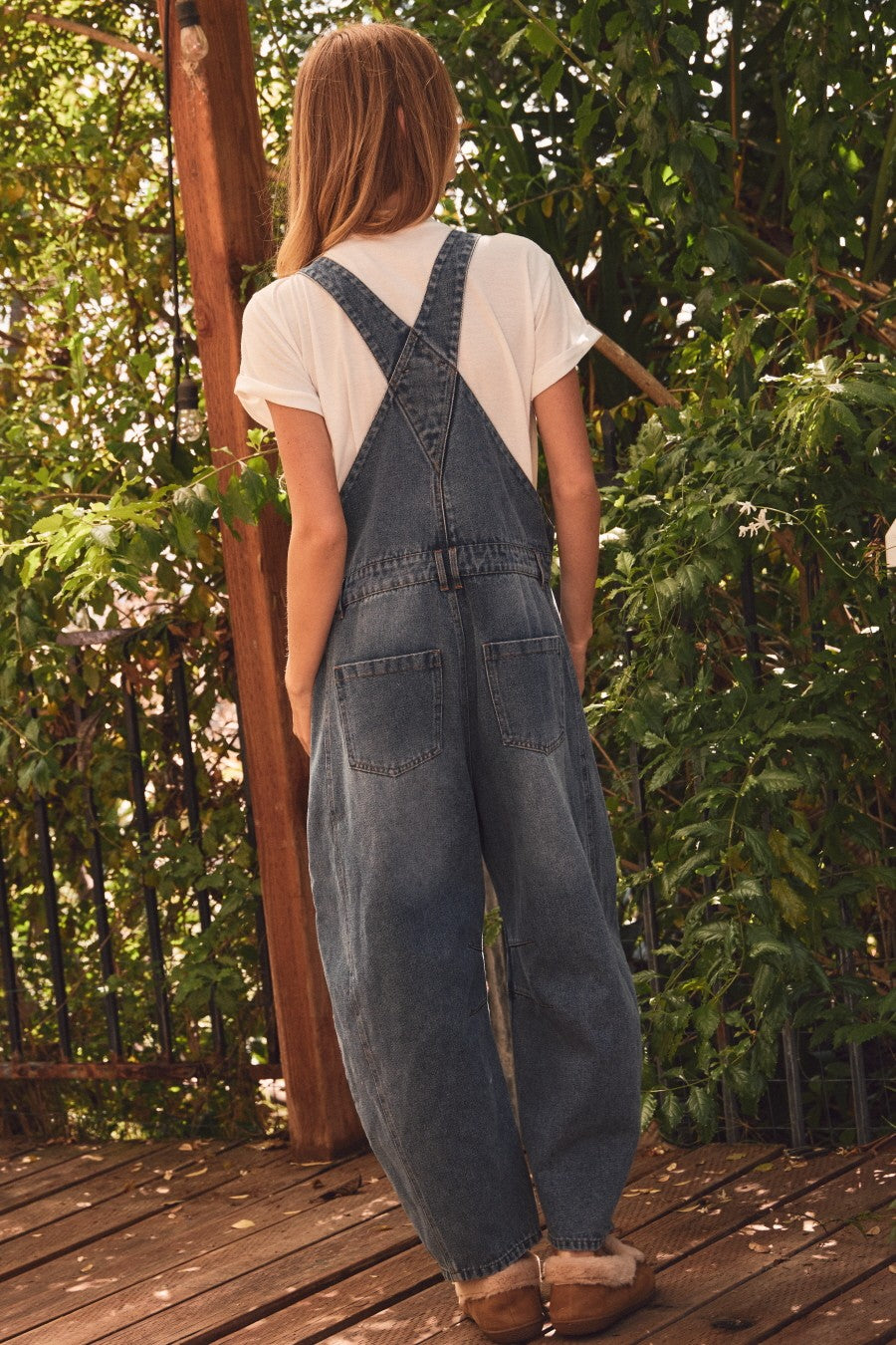 woman wearing a pair of denim overalls with a white t-shirt underneath, standing against a leafy background