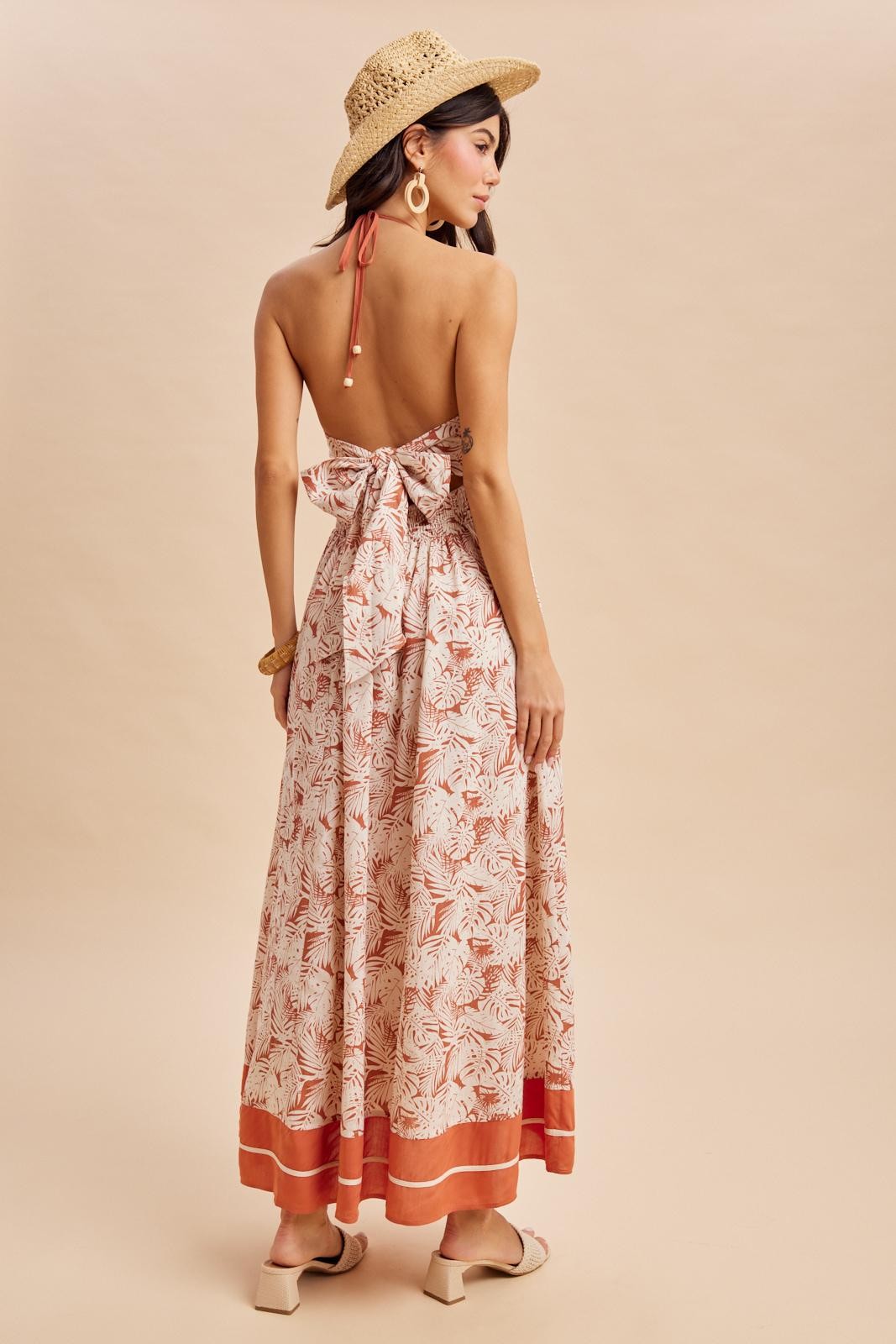 woman wearing a backless floral maxi dress with a bow detail and a wide-brimmed hat, standing against a neutral background