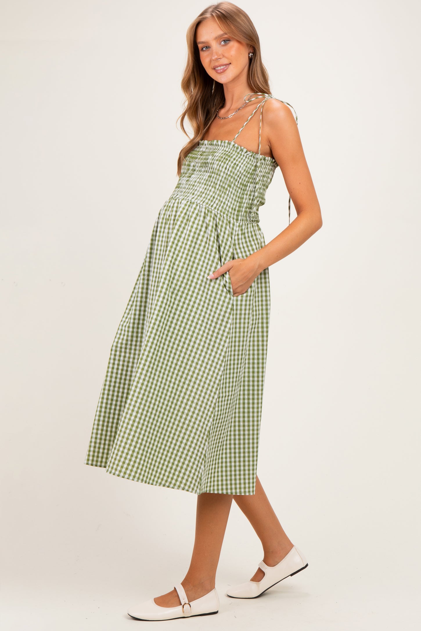 woman wearing a green and white gingham maternity dress with a smocked bodice and pockets, showing a baby bump