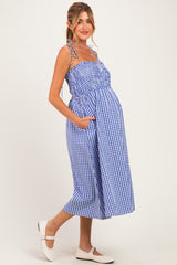 woman wearing a blue and white gingham maternity dress with adjustable straps and side pockets, showcasing her baby bump