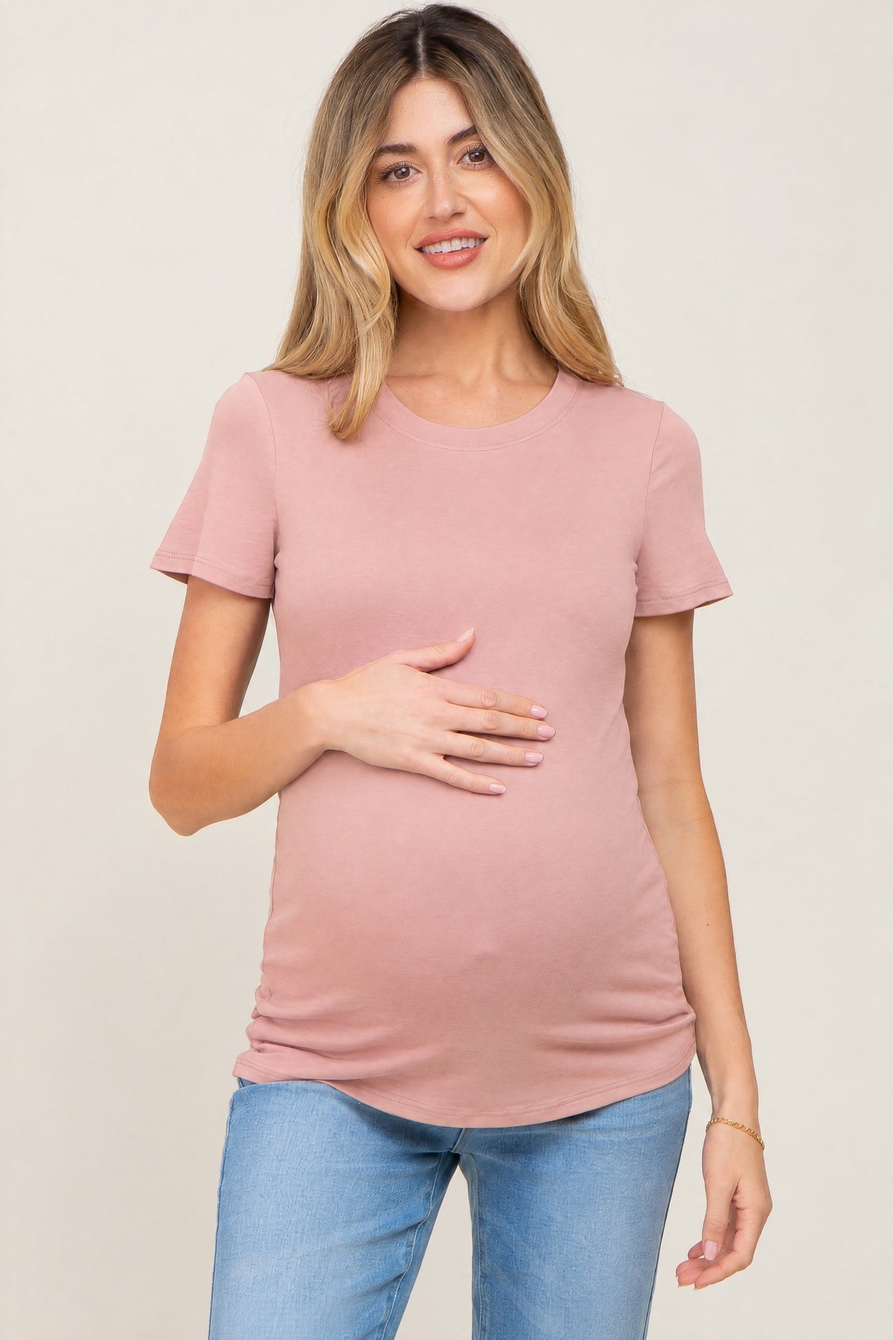 woman wearing a dusty rose fitted maternity top and holding her baby bump, paired with light blue jeans