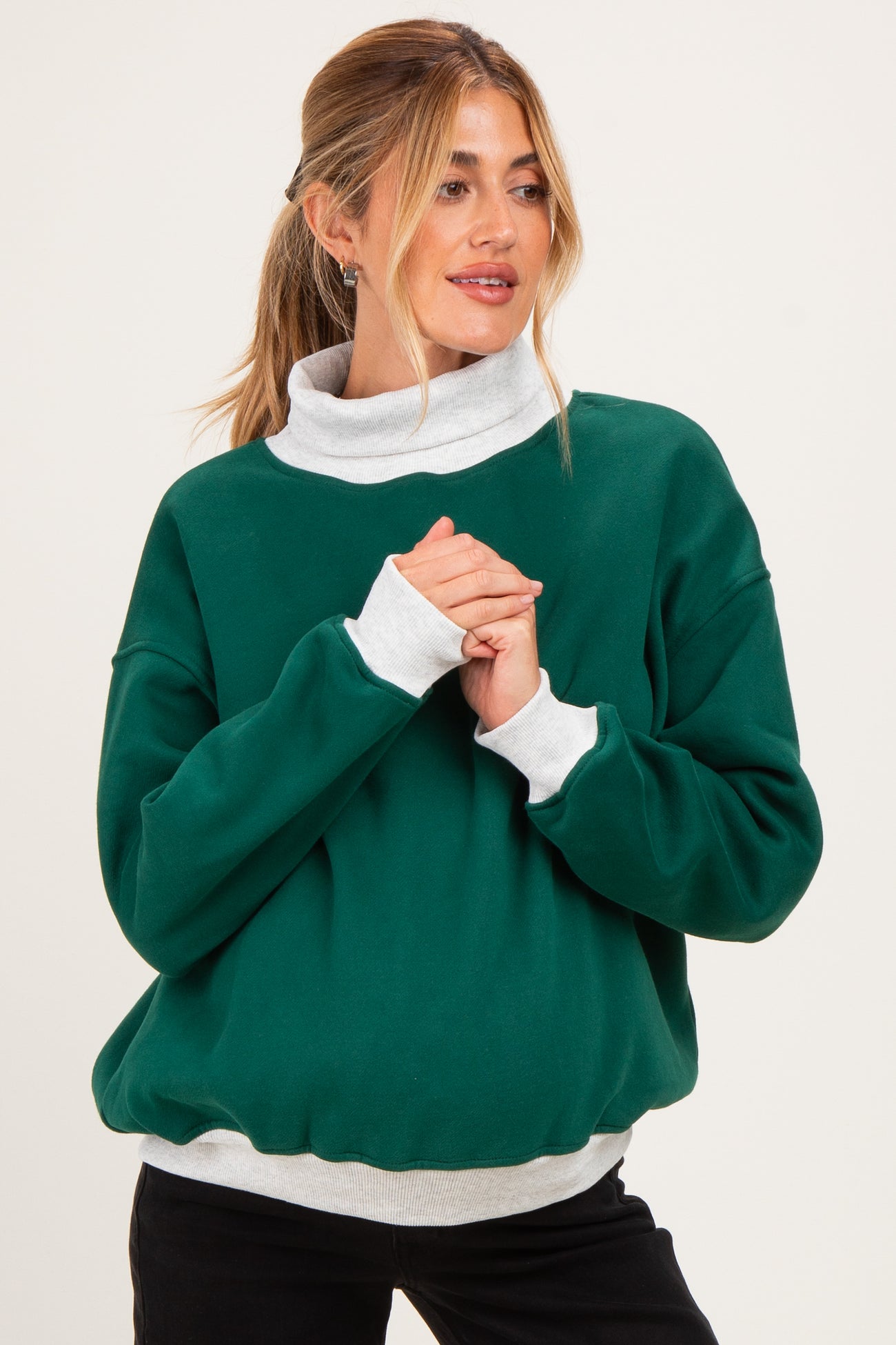 woman wearing a green maternity sweatshirt with a light gray collar and cuffs, standing with hands clasped in front